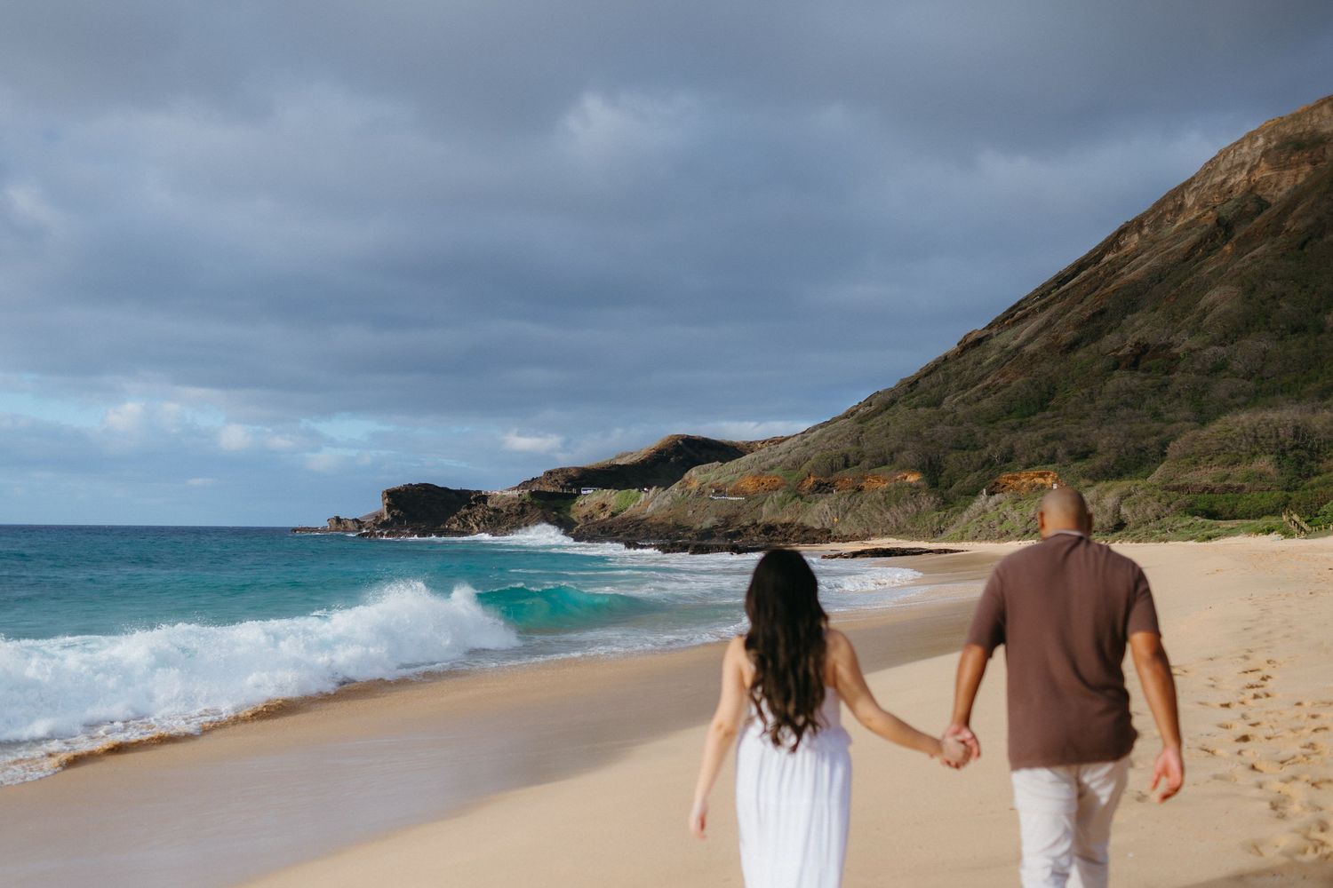 Couple holding hands walking along a sandy beach with turquoise waves and dramatic cliffs in Hawaii.
