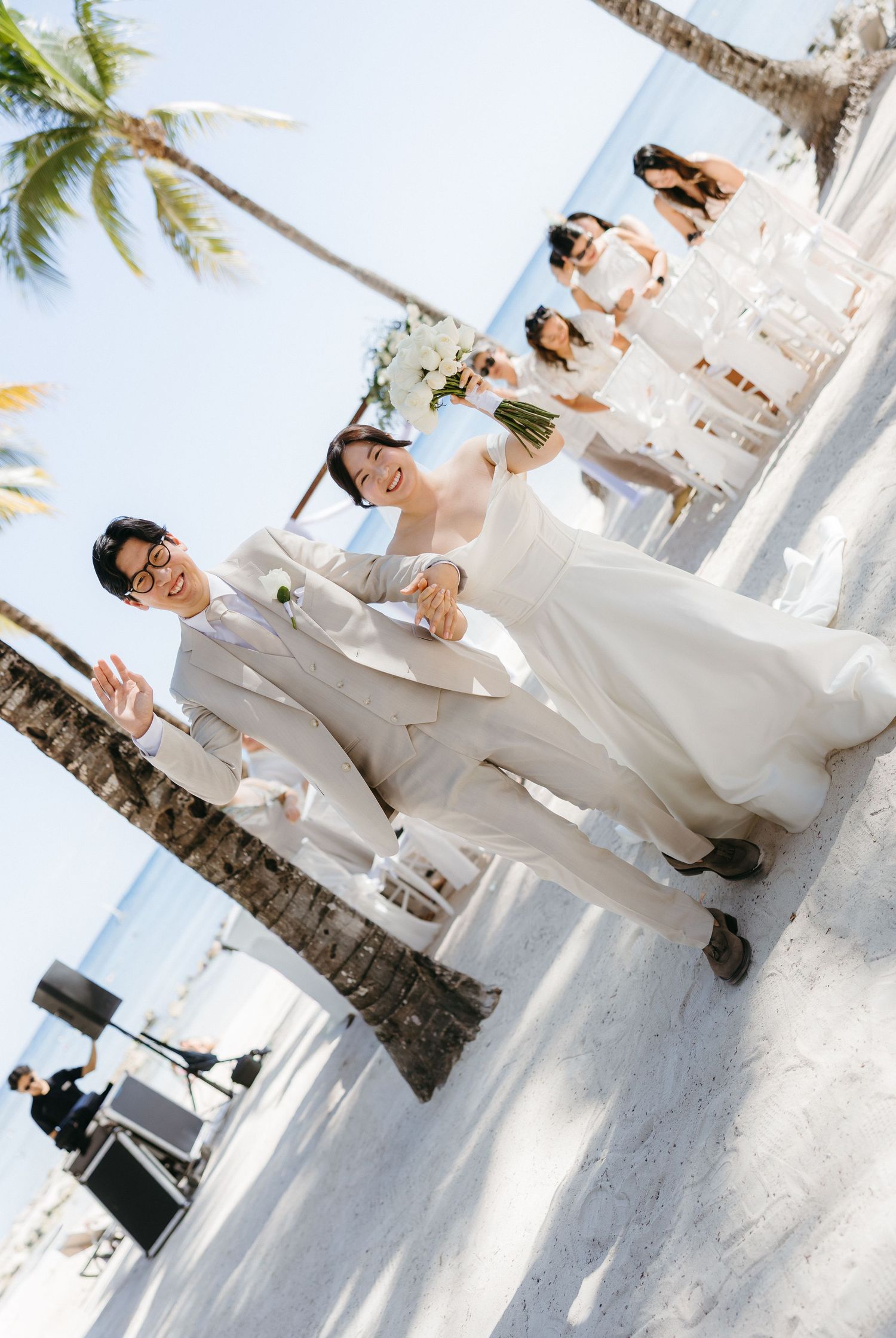 Smiling newlyweds walking down beach wedding aisle with bridesmaids and ocean backdrop.