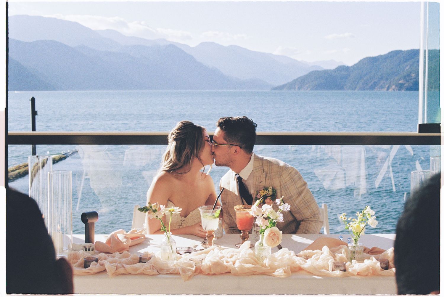 Bride and groom sharing a kiss at a sweetheart table overlooking a stunning mountain lake at their wedding reception.