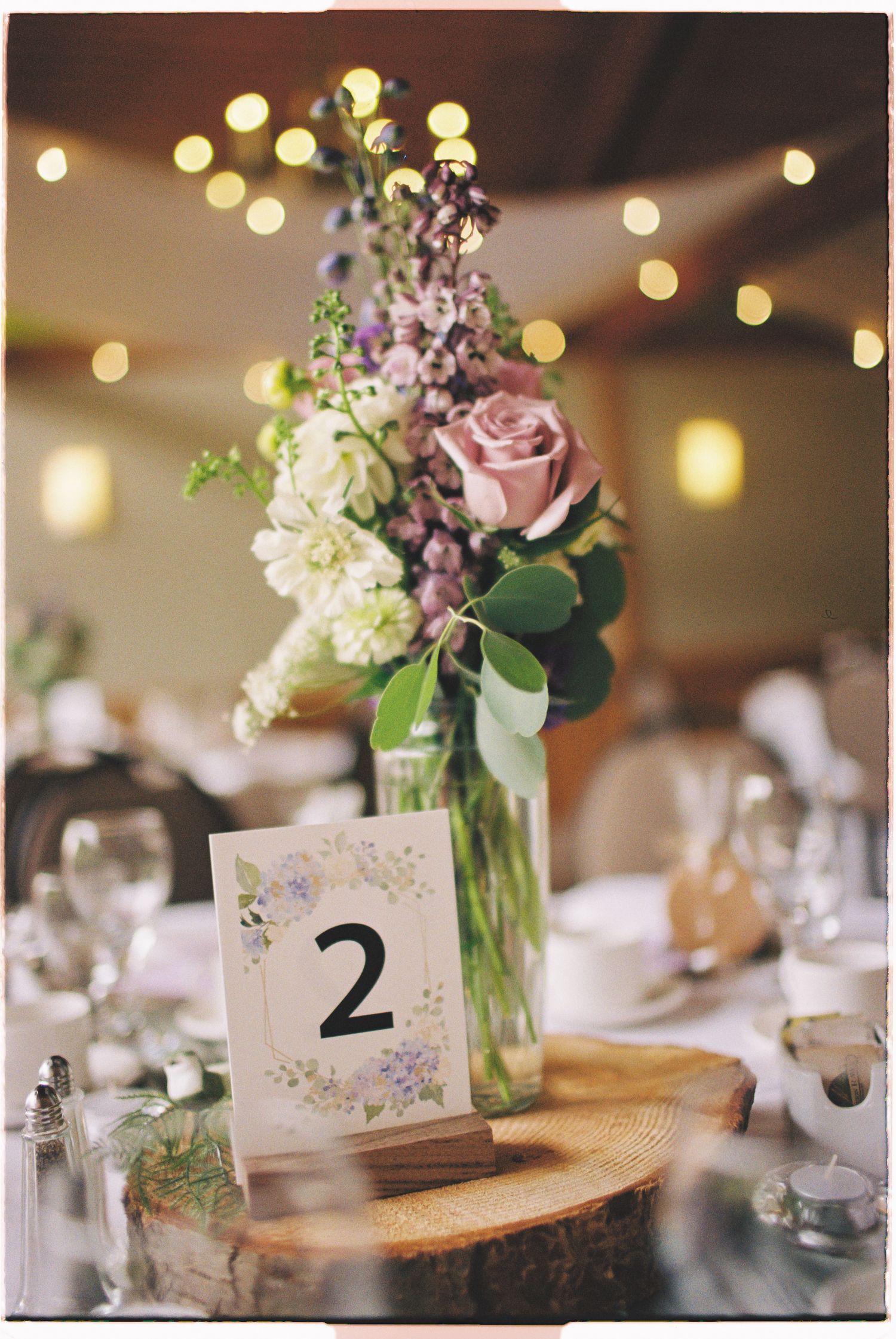 Elegant wedding table centerpiece with purple and white flowers in a glass vase beside a floral table number sign.
