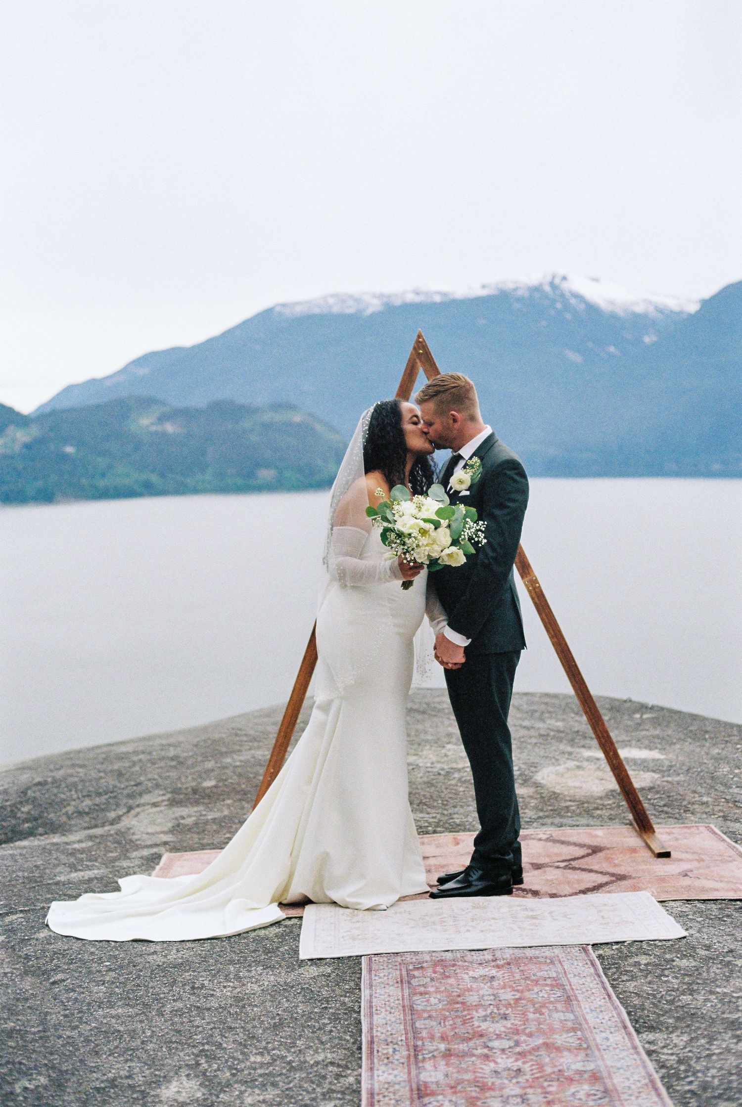 Bride and groom kissing under a wooden triangular arch with mountain lake backdrop at an outdoor wedding ceremony.