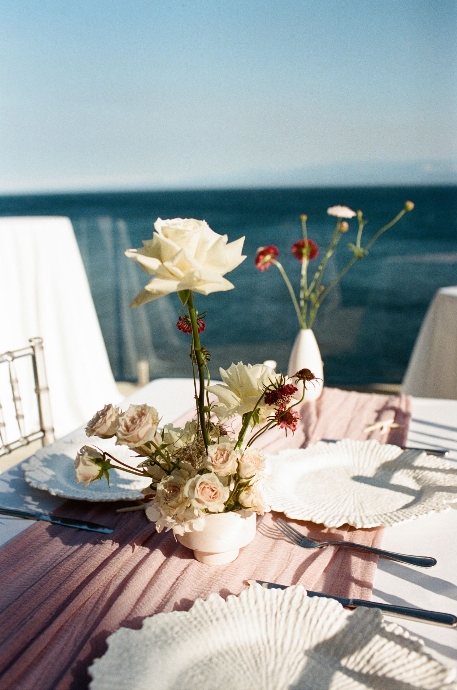 Elegant outdoor wedding table setting with white roses and wildflowers overlooking a calm blue ocean.