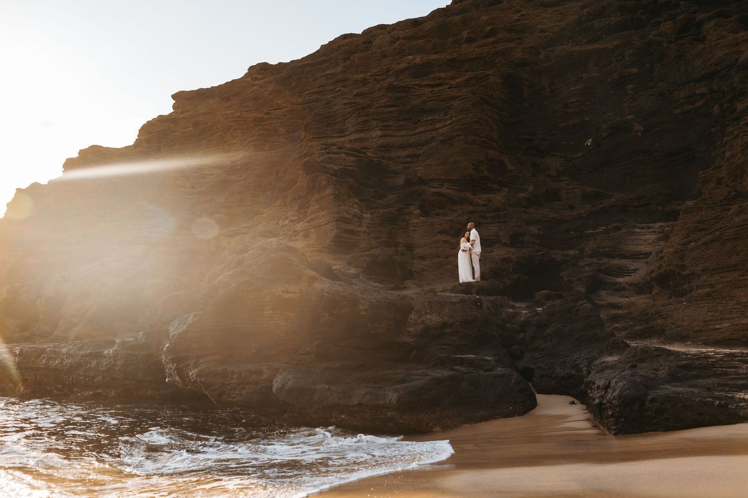 Couple in white embracing on dramatic rocky coastal cliffs at sunset with waves crashing below.