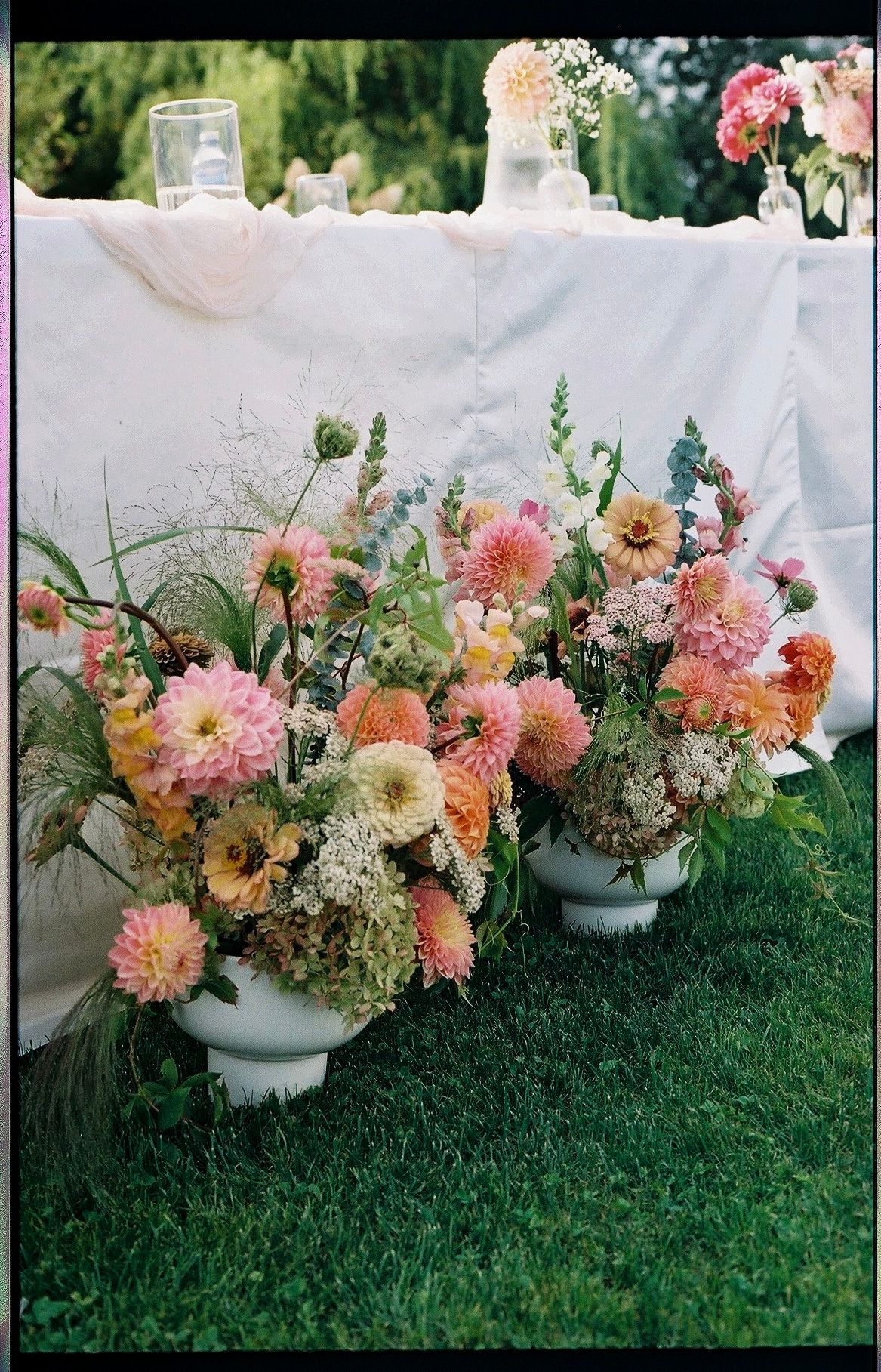Two white urns overflowing with lush pink, peach, and coral dahlia floral arrangements on grass at an outdoor wedding reception.