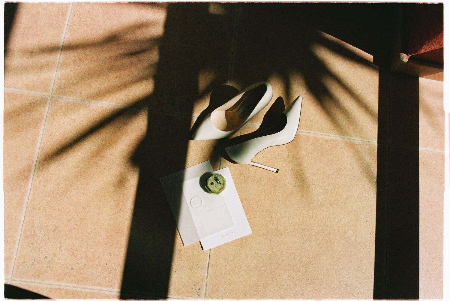 White stiletto bridal shoes and wedding accessories with tropical palm shadows on tile.