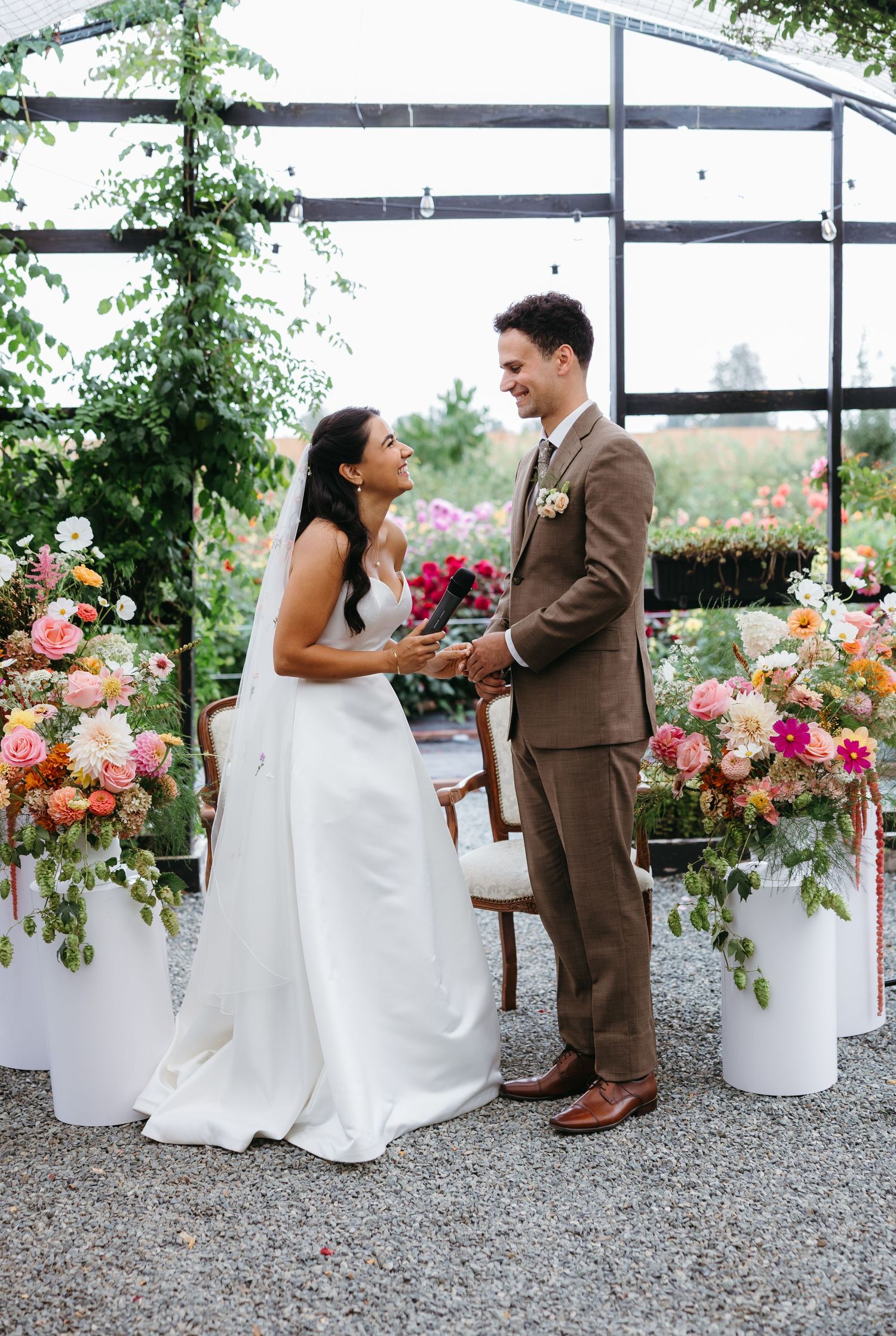 Bride and groom exchange vows in a flower-filled greenhouse surrounded by colorful pink and orange floral arrangements.