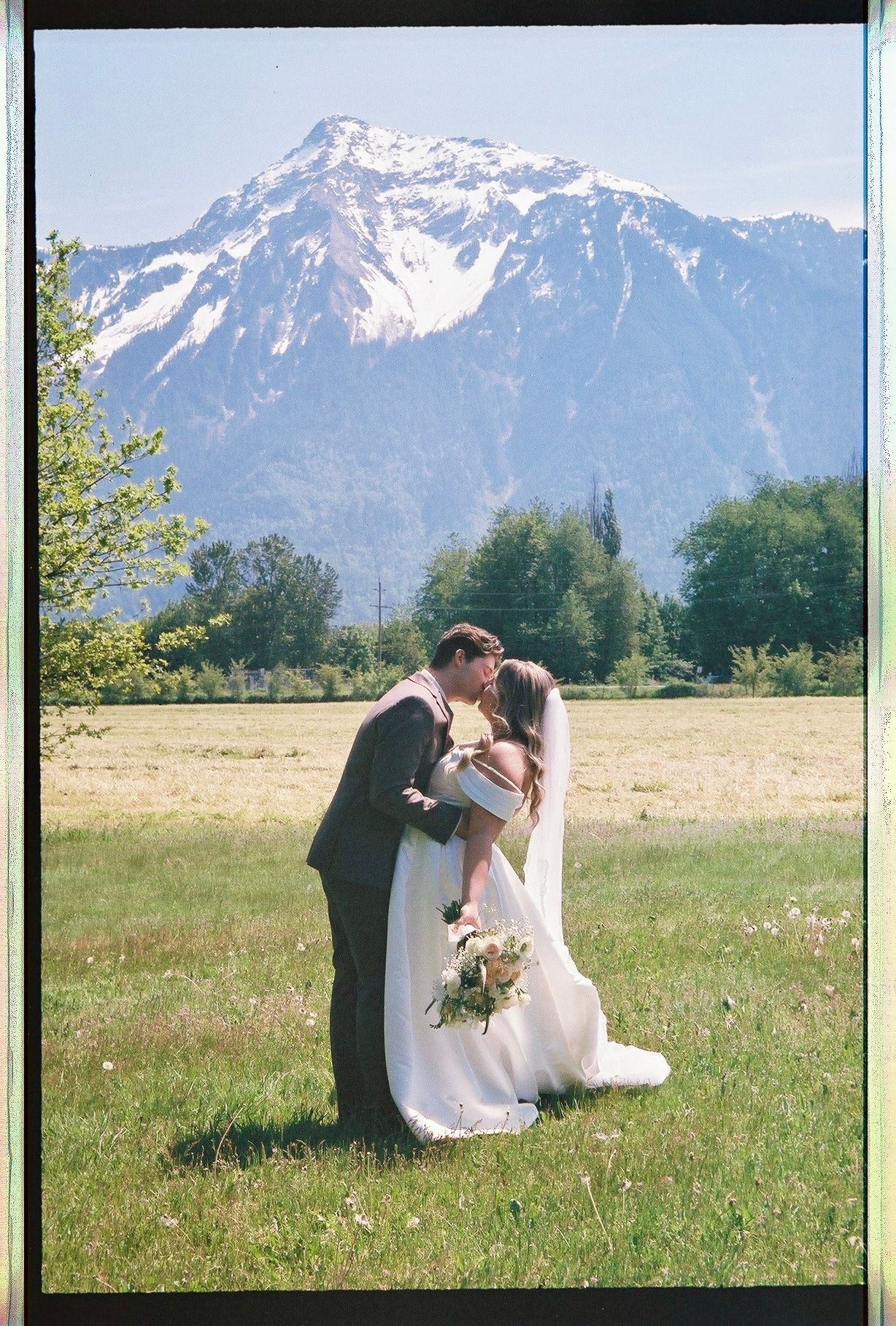 Bride and groom kissing in a green meadow with a snow-capped mountain in the background.