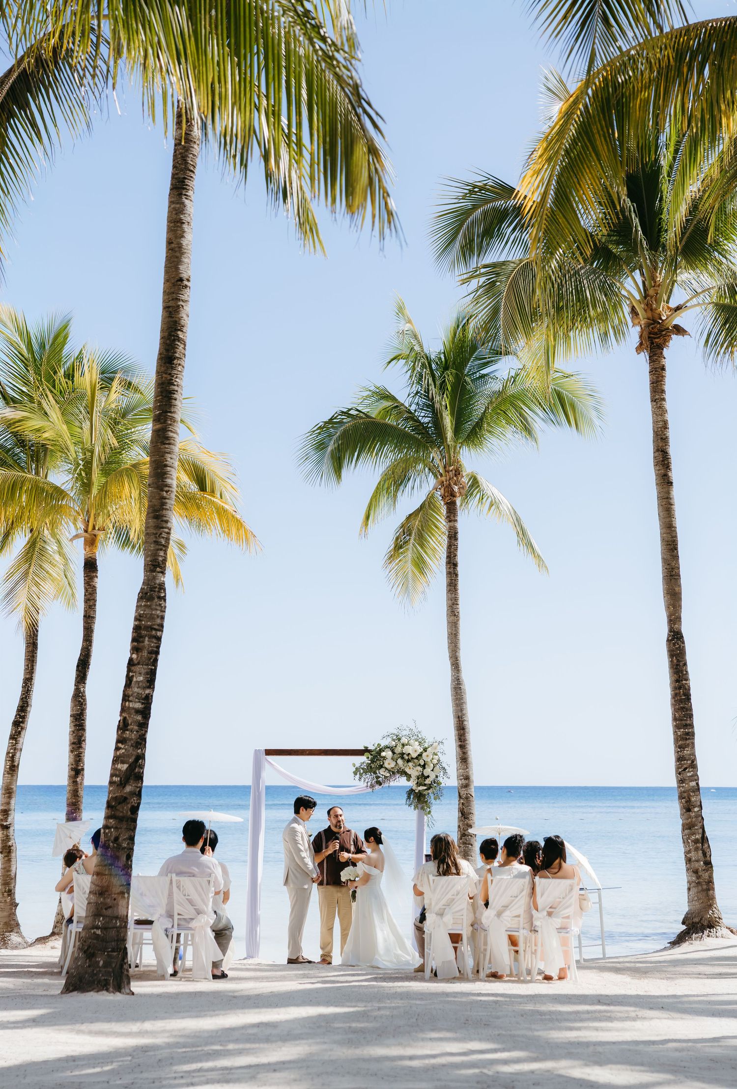 Intimate beach wedding ceremony under tall palm trees with ocean backdrop in tropical destination.