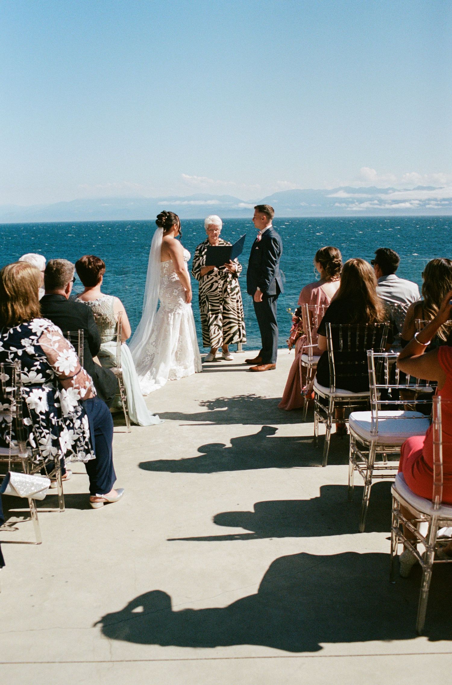 Outdoor wedding ceremony by the ocean with bride, groom, and officiant at the altar while guests watch from chairs.