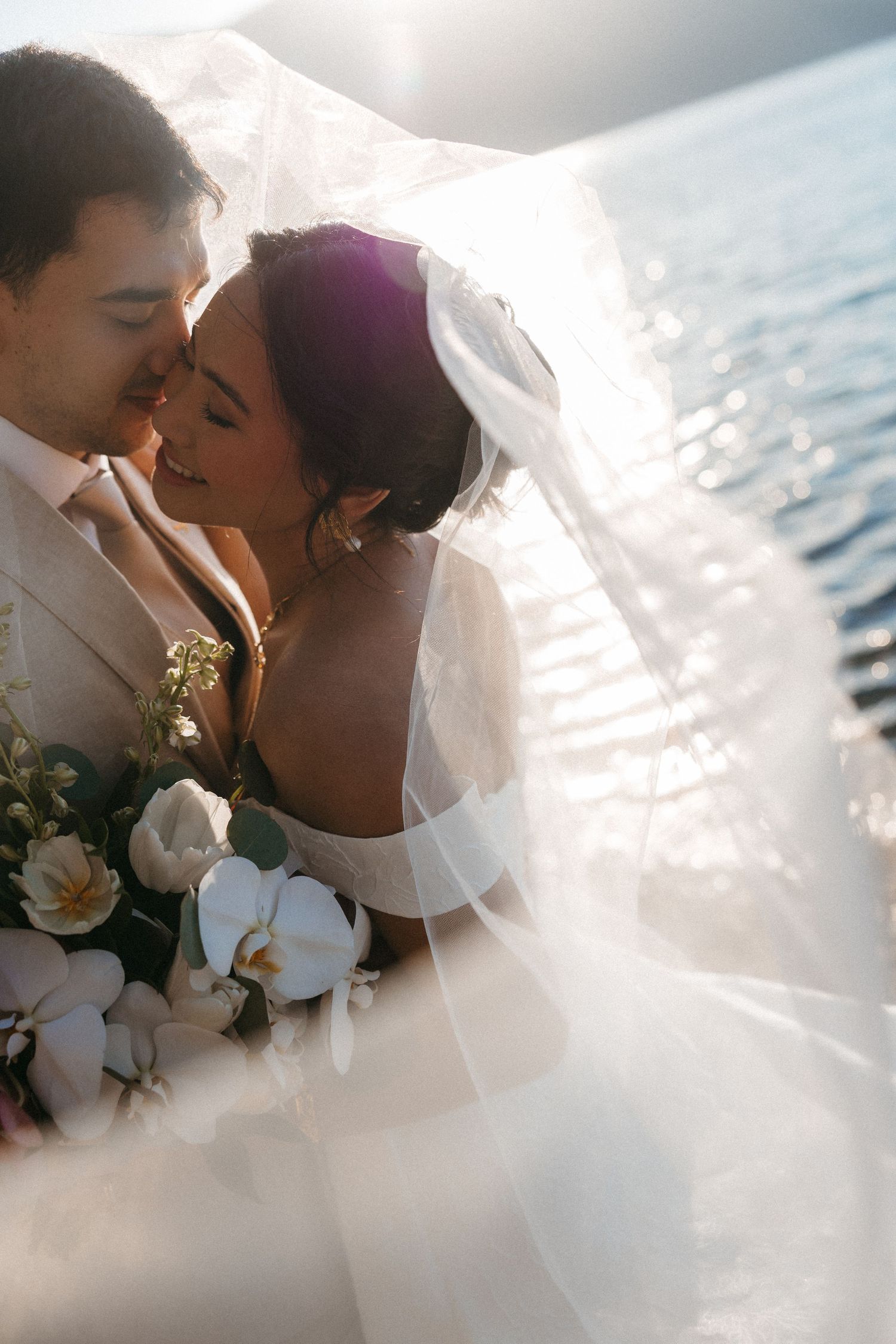Bride and groom share romantic moment under flowing veil by sparkling water, bride holding white orchid bouquet.