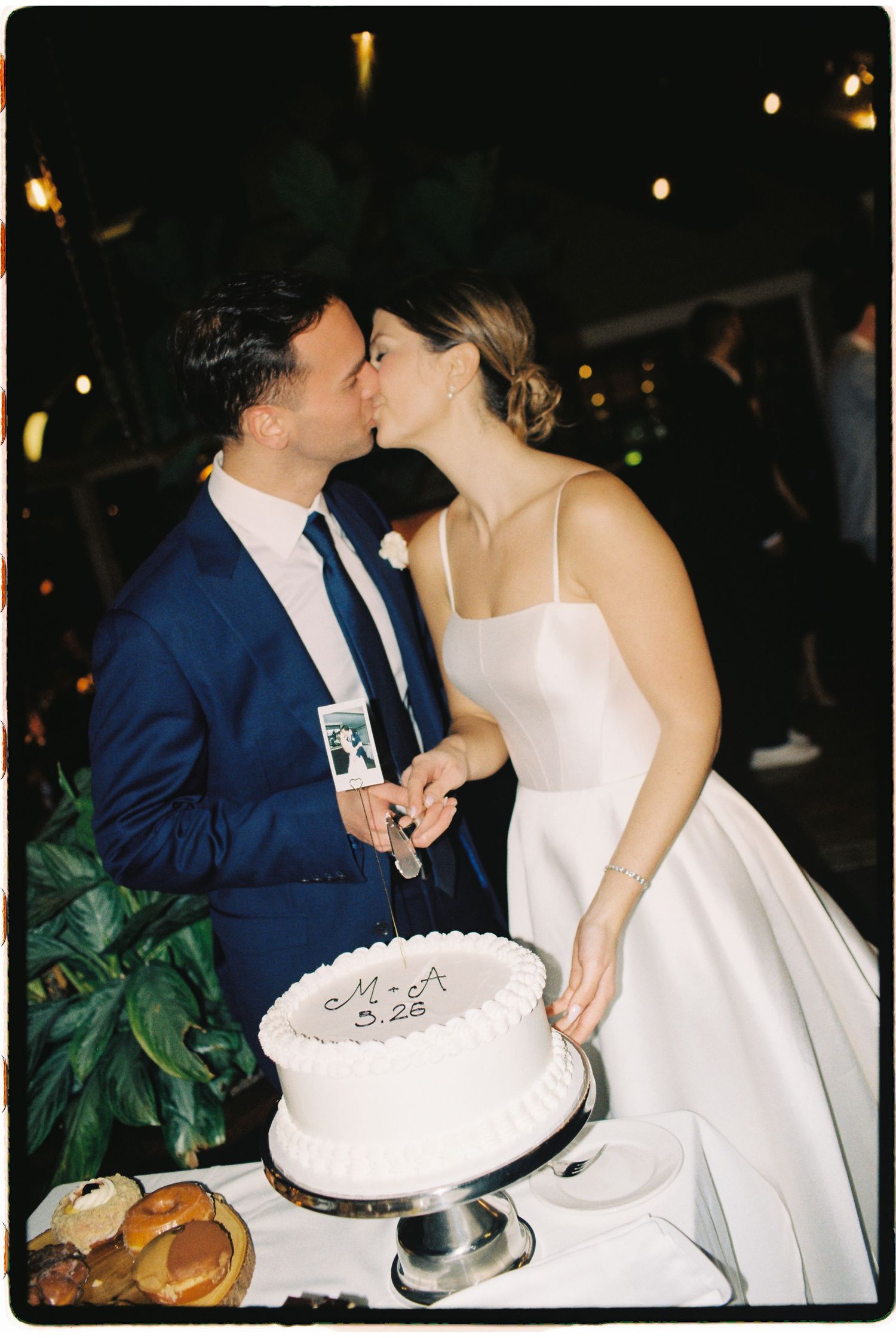 Newlyweds kissing while cutting their white wedding cake at a nighttime reception.