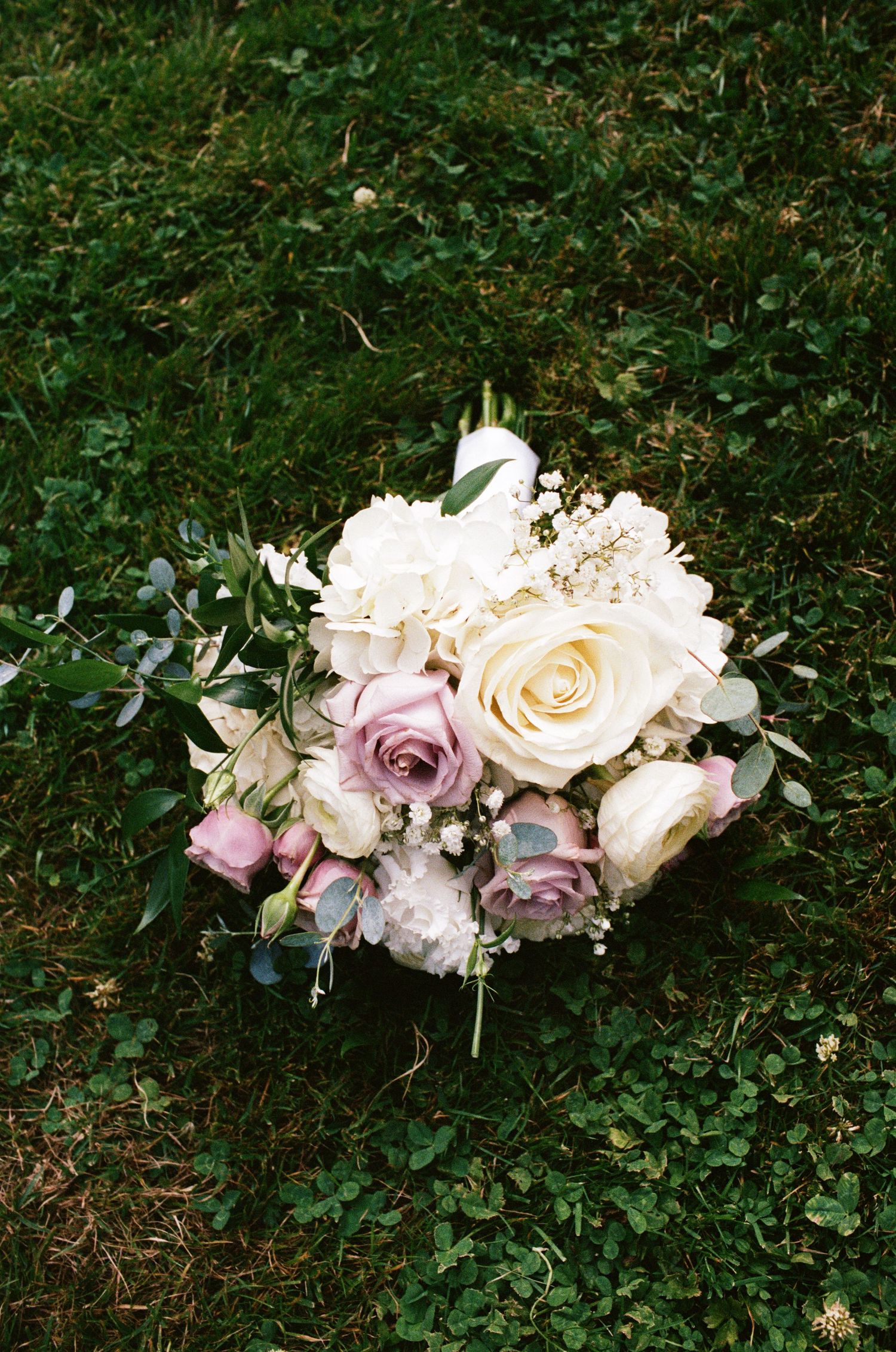 Romantic bridal bouquet of white hydrangeas, cream and mauve roses resting on lush green grass.