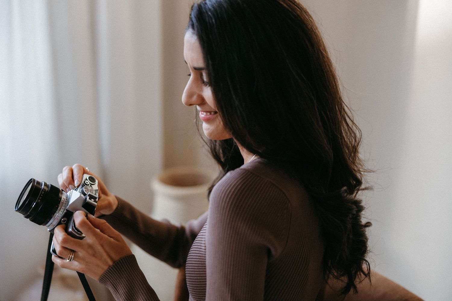 A smiling young woman with long dark hair holds a vintage camera, wearing a brown ribbed sweater in a bright room.