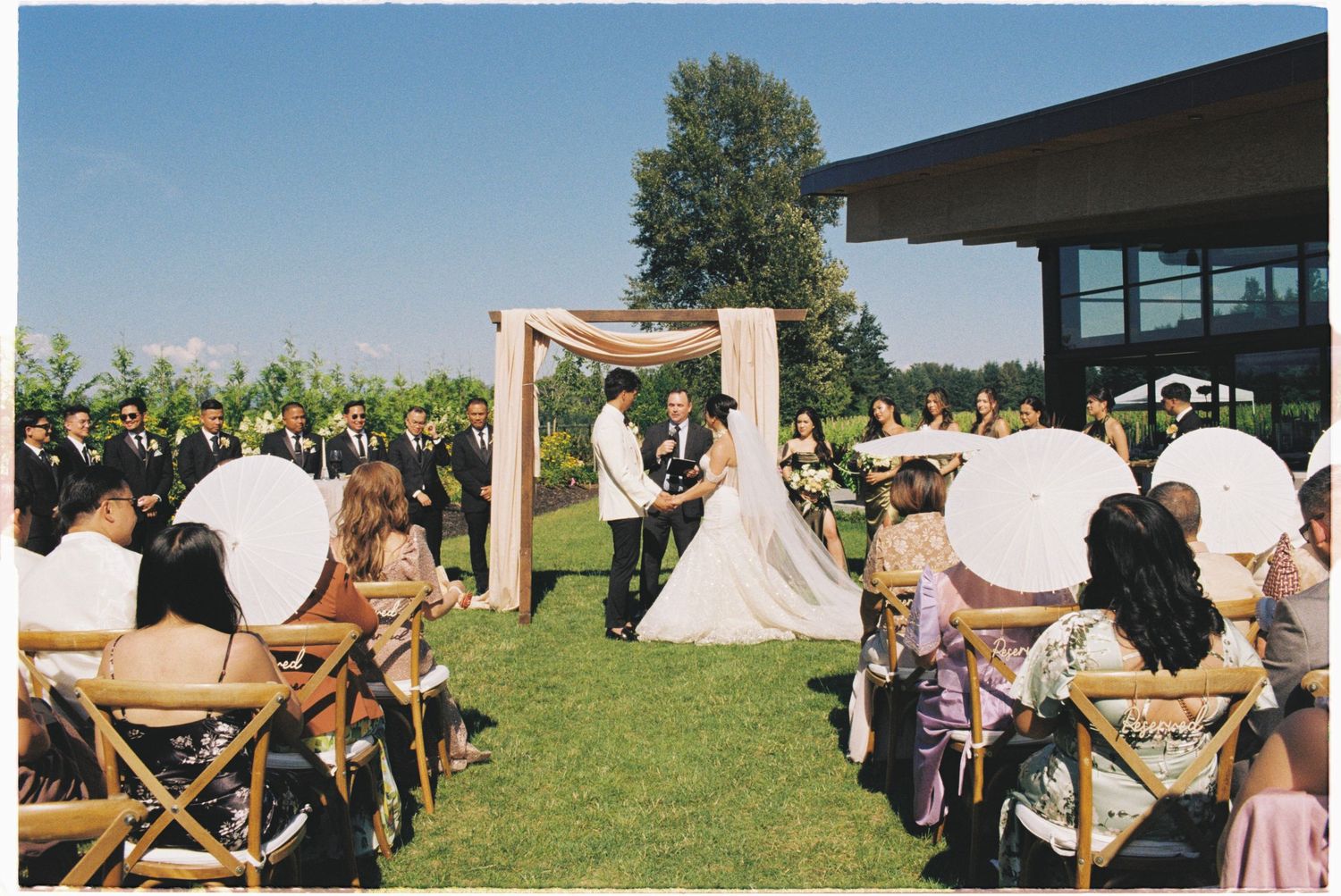 Outdoor wedding ceremony on green lawn with guests seated on wooden chairs under a floral arch.