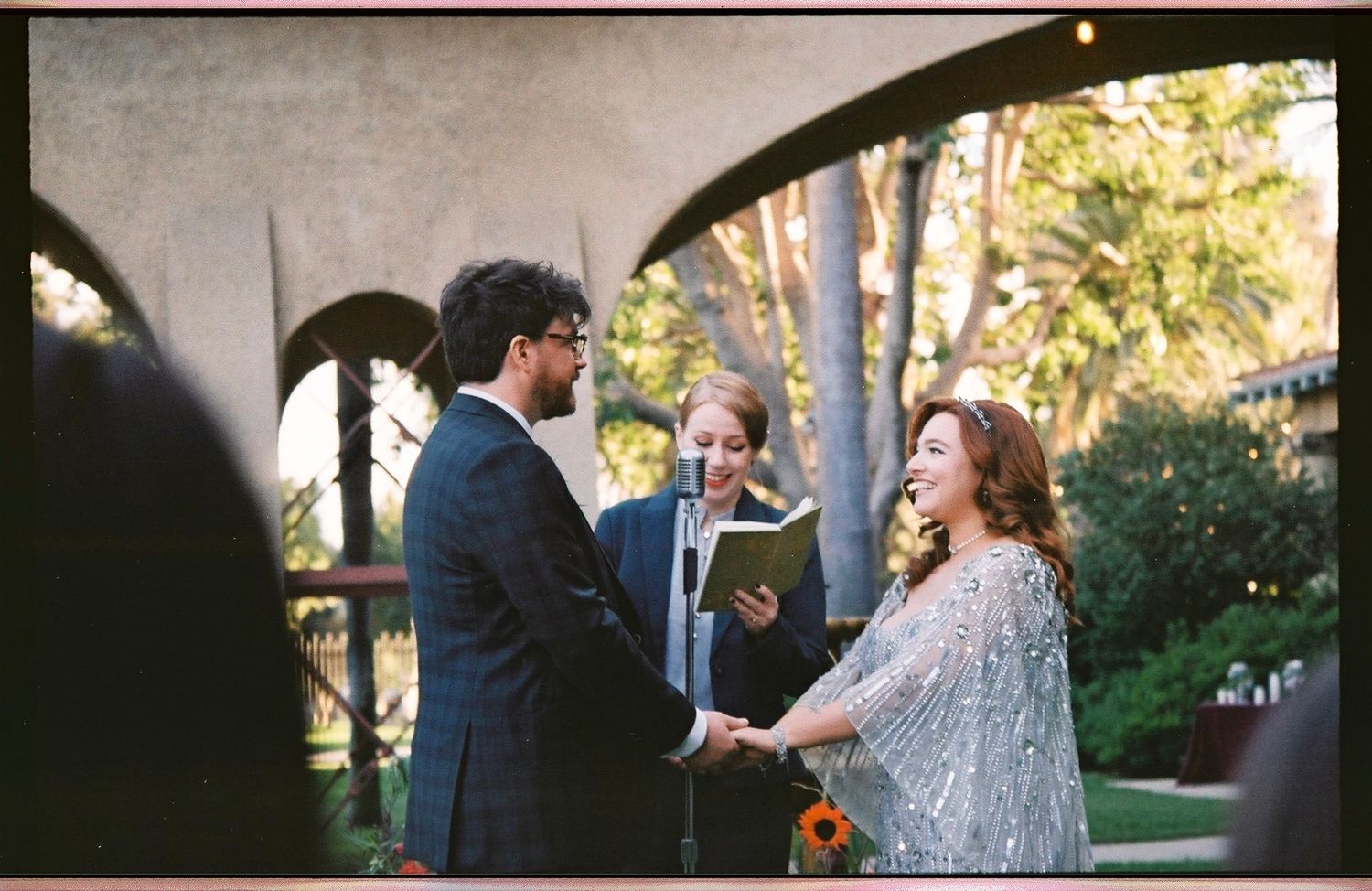 Bride in silver gown holding groom's hands during intimate outdoor wedding ceremony with officiant.