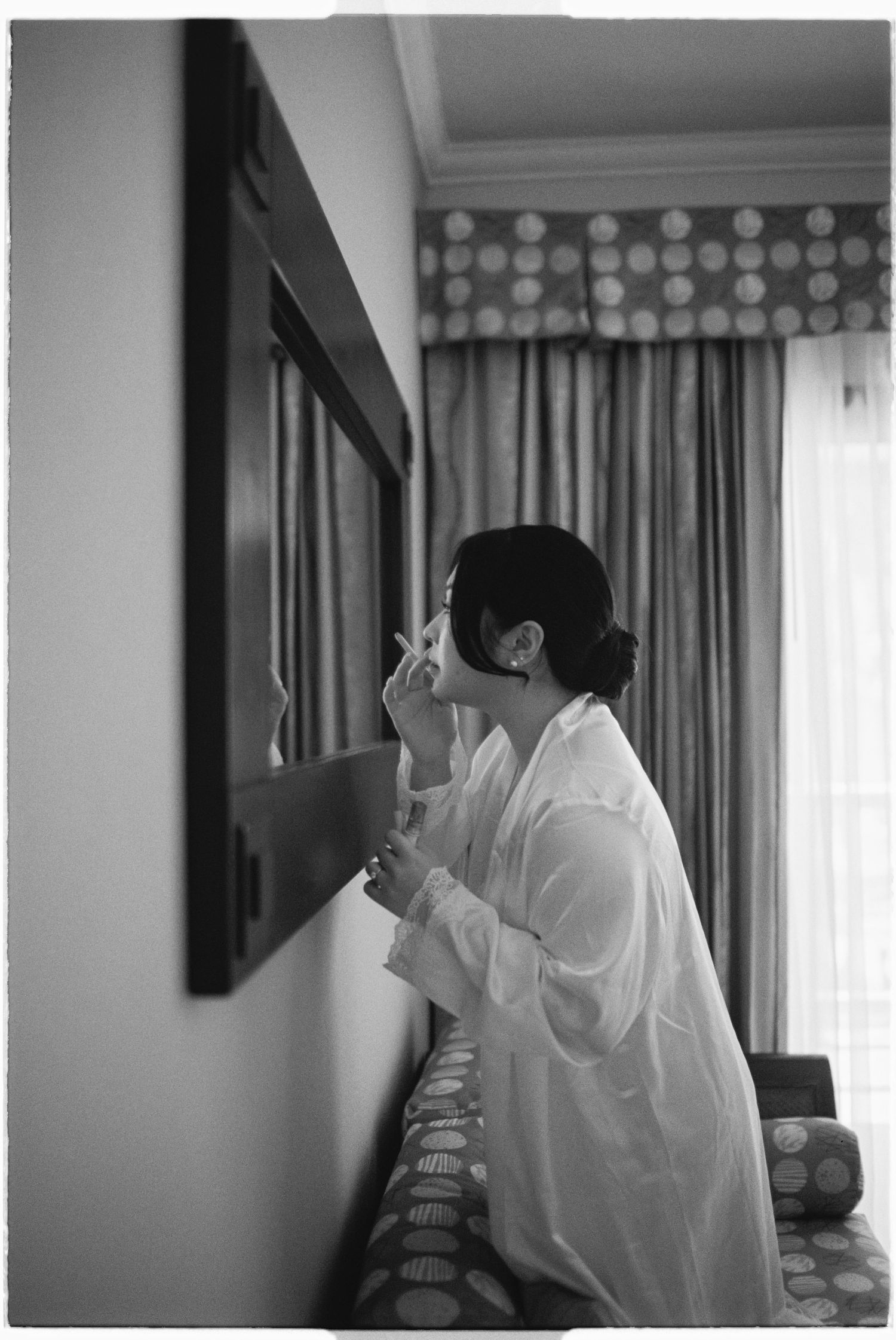 Bride in white robe applying makeup at mirror in black and white hotel room photo.