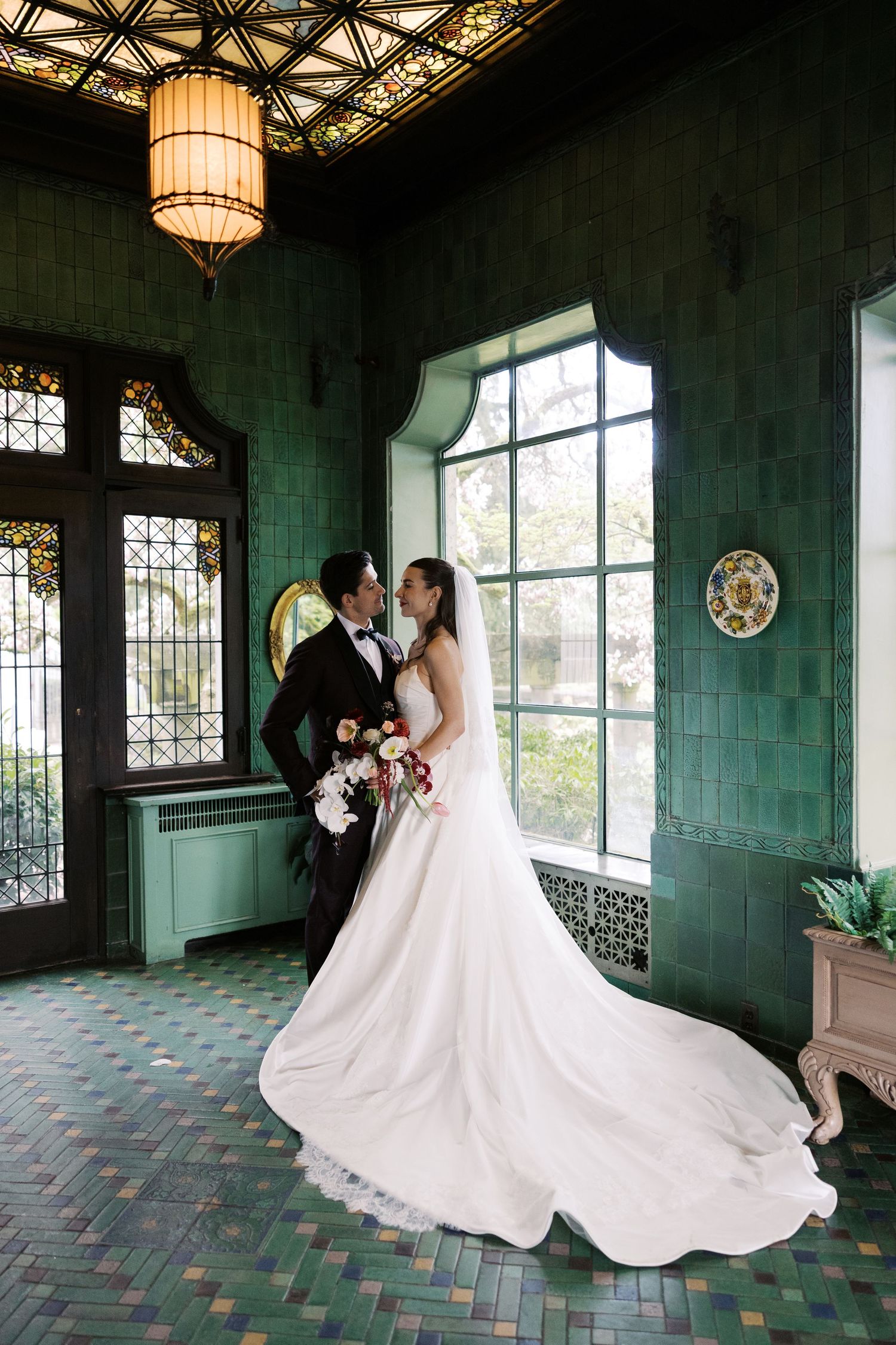 Bride in elegant gown with long veil and groom in tuxedo posing in green tiled Art Nouveau room.