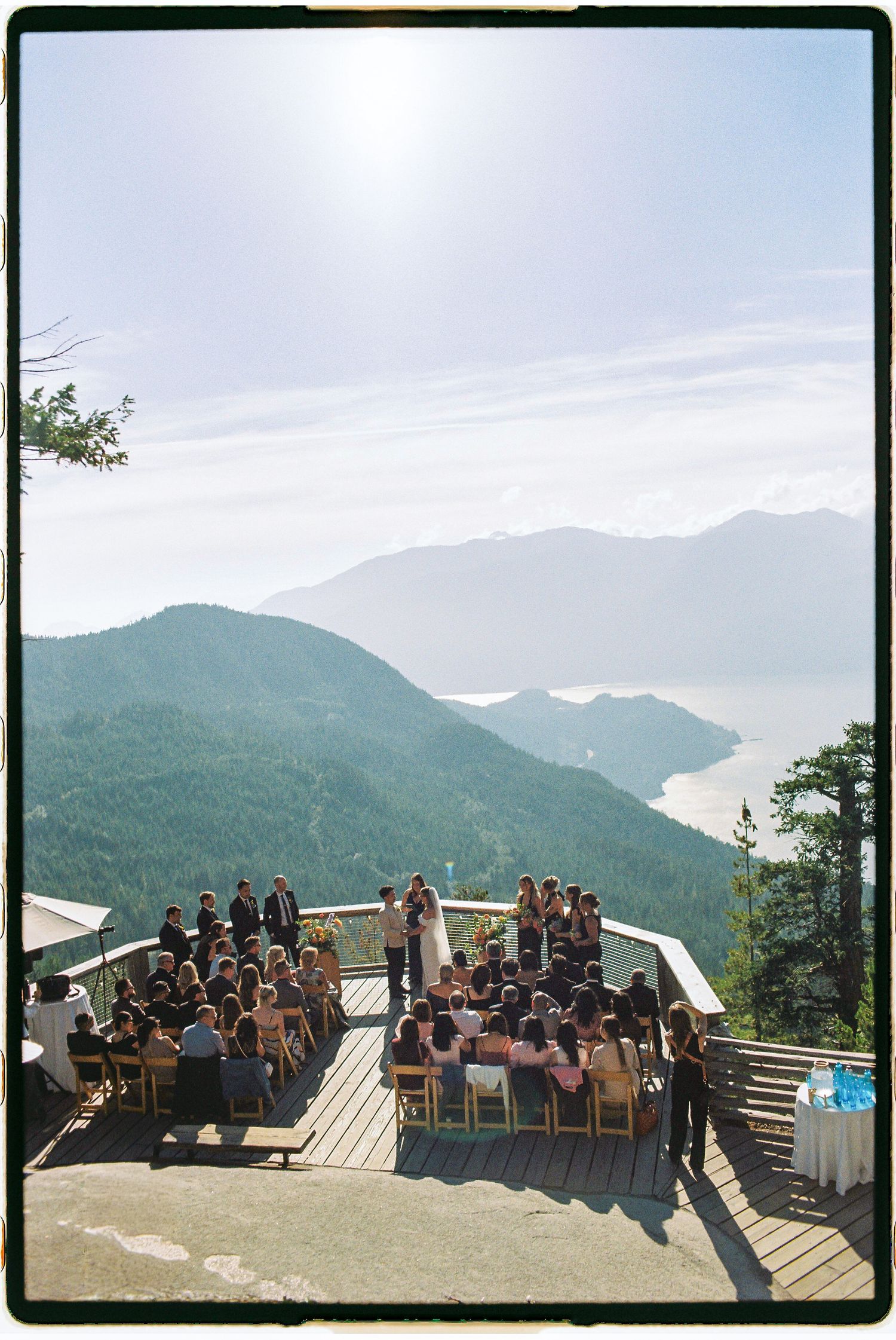 Aerial view of an outdoor mountain top wedding ceremony on a deck overlooking stunning green mountains and ocean inlet.