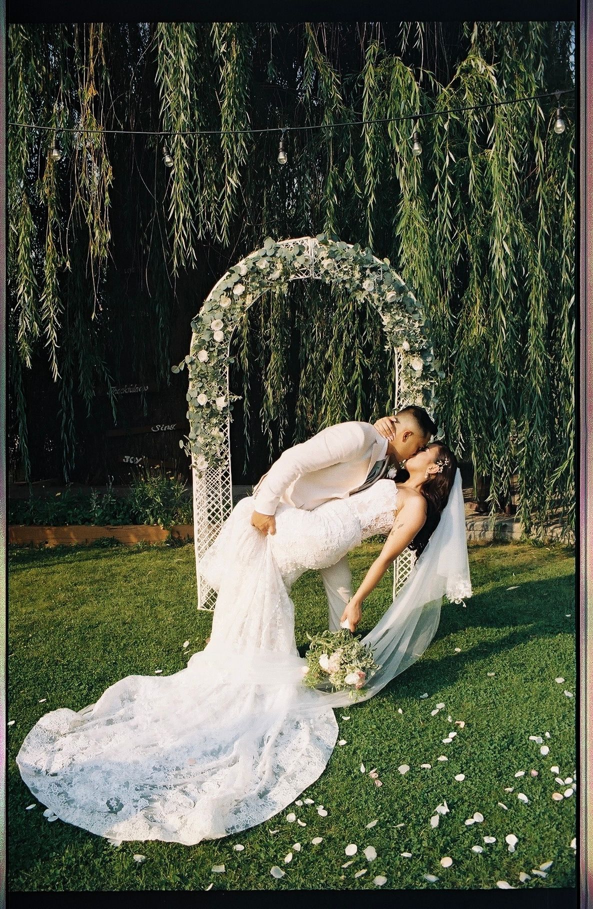 Bride and groom sharing a romantic dip kiss under a floral arch with weeping willow trees.