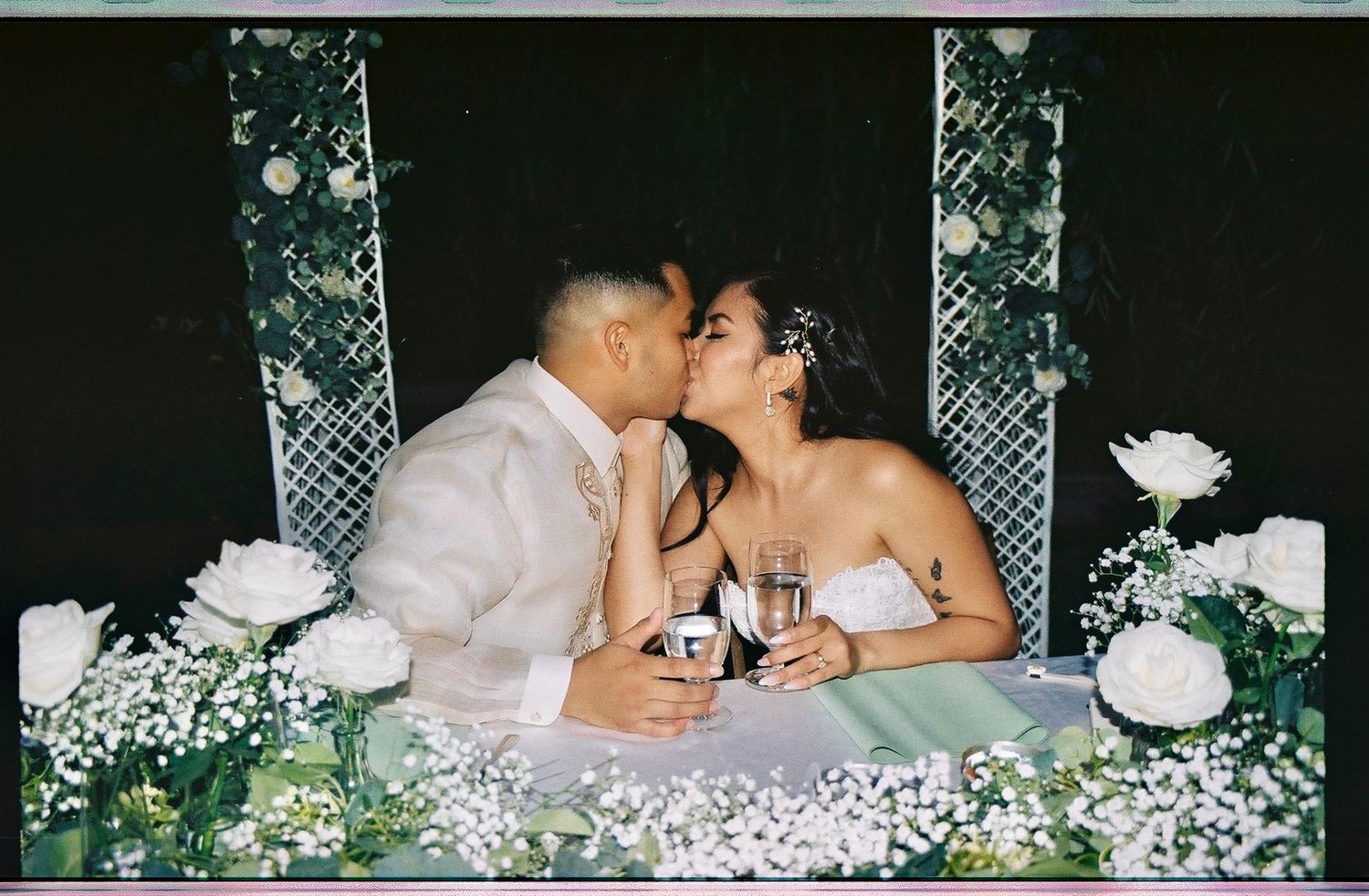 Newlyweds sharing a kiss at sweetheart table adorned with white roses and baby's breath at reception.