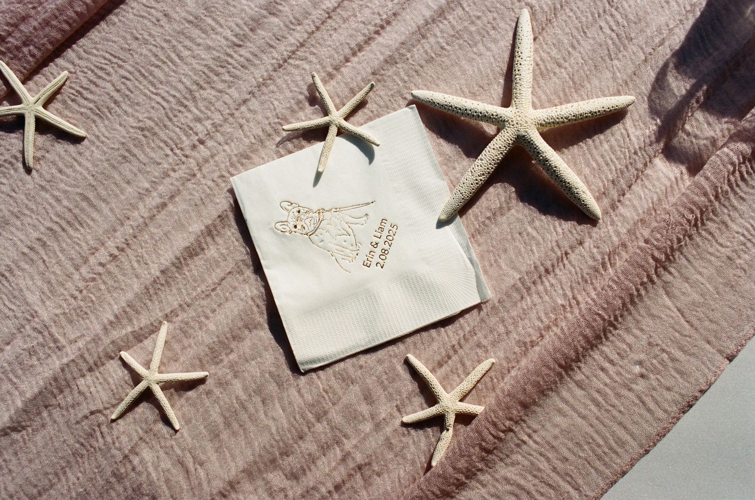 Starfish arranged around a small white gift tag on pink linen fabric in natural sunlight.