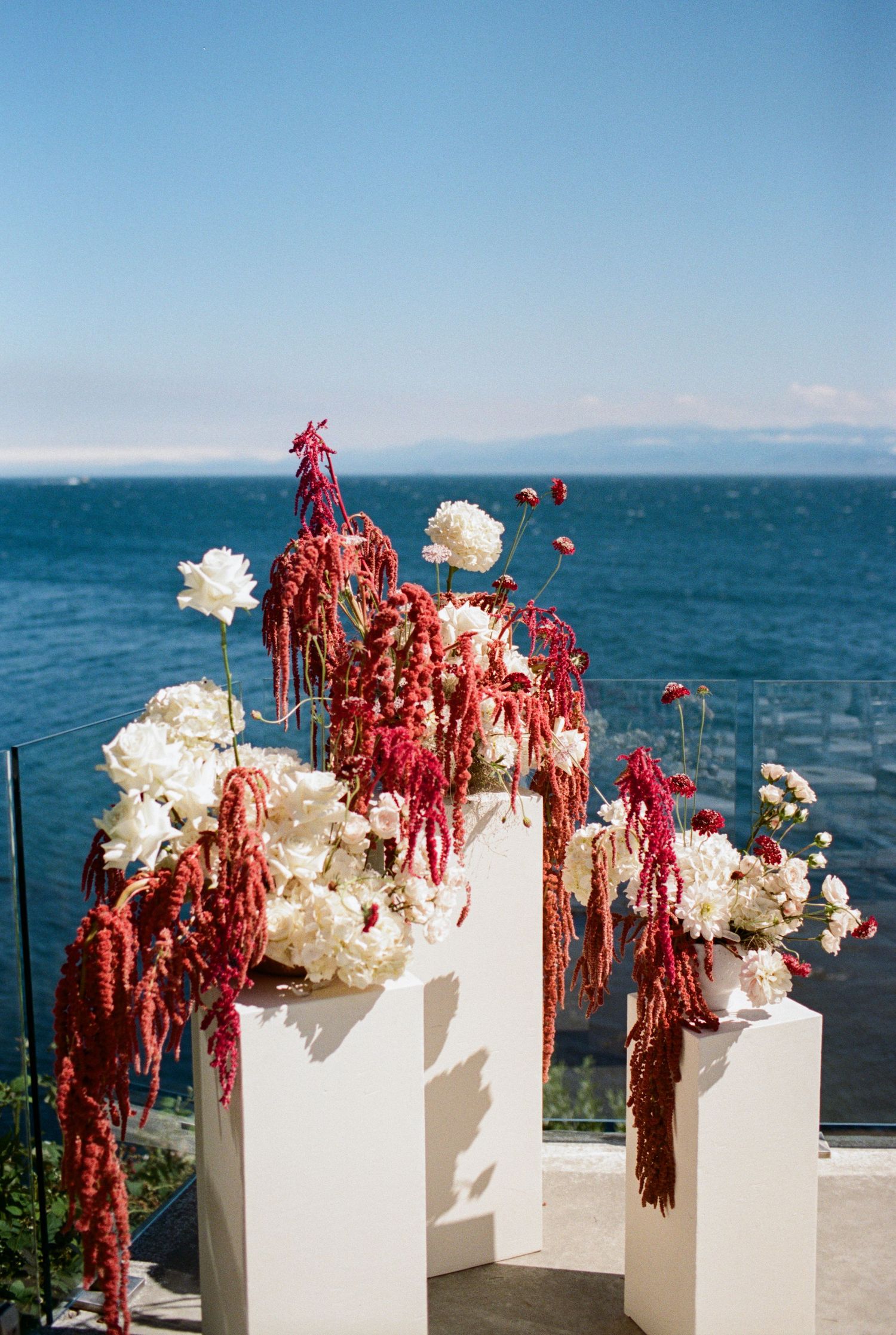 Dramatic wedding ceremony floral arrangement with red amaranthus and white peonies against blue ocean sky.