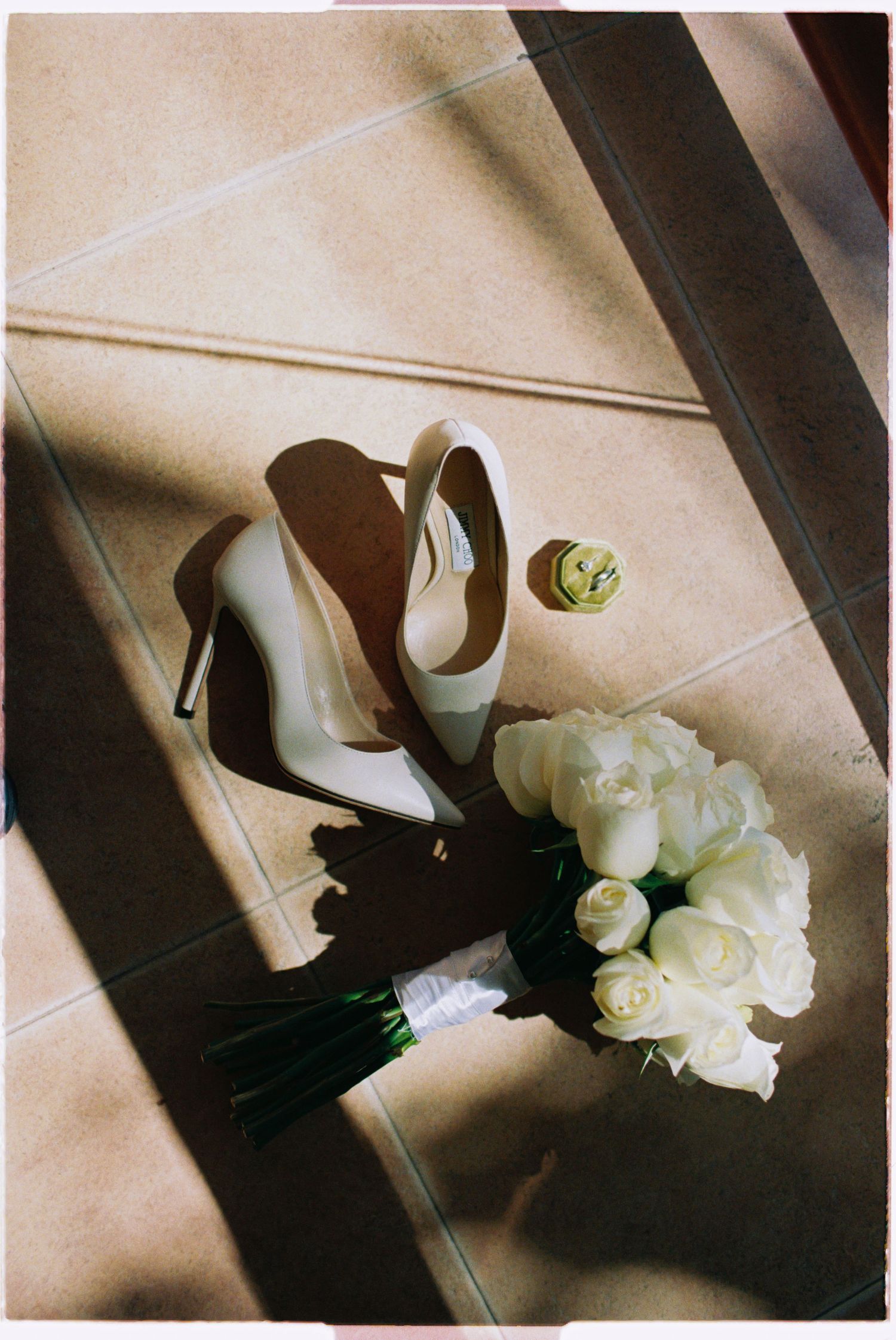 White bridal heels, bouquet of white roses, and perfume arranged on sunlit tile floor.