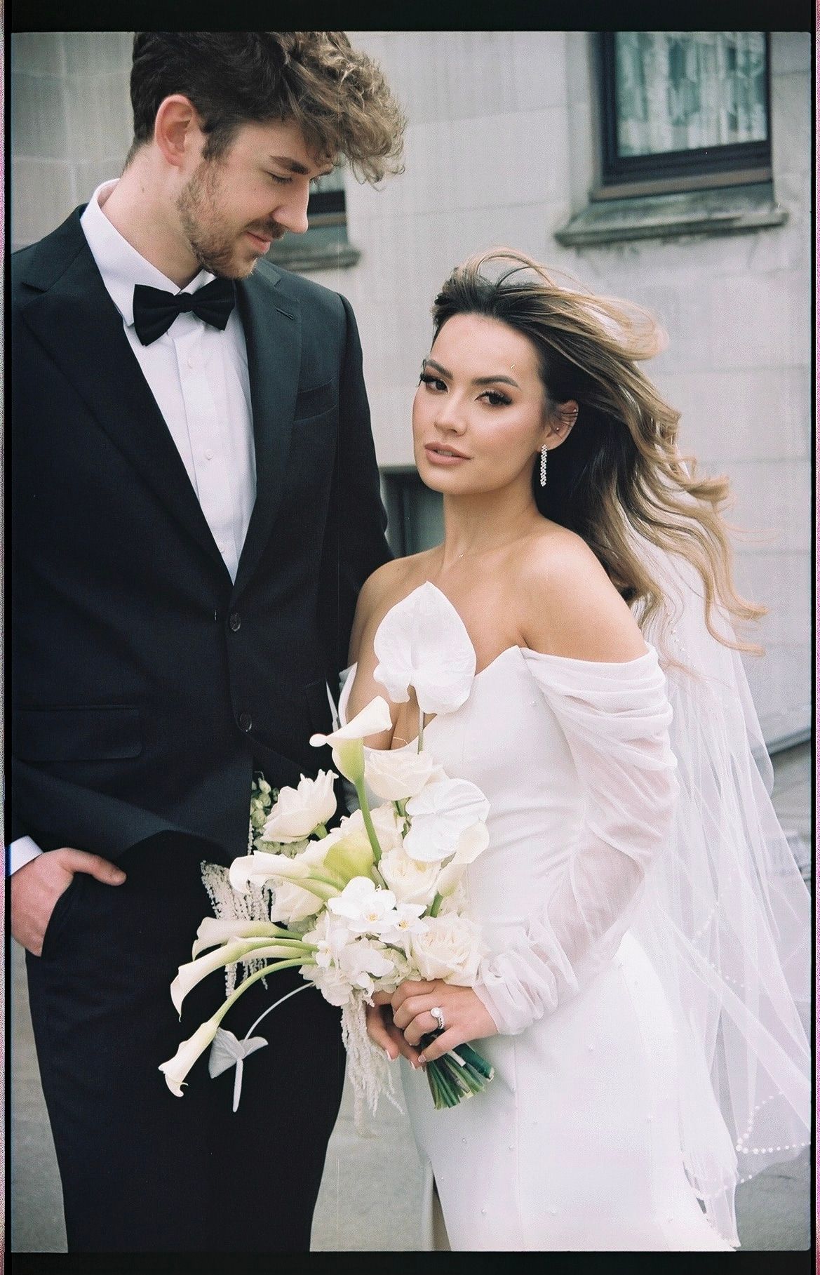 Elegant bride in off-shoulder white gown holding white tulip bouquet stands with groom in black tuxedo.