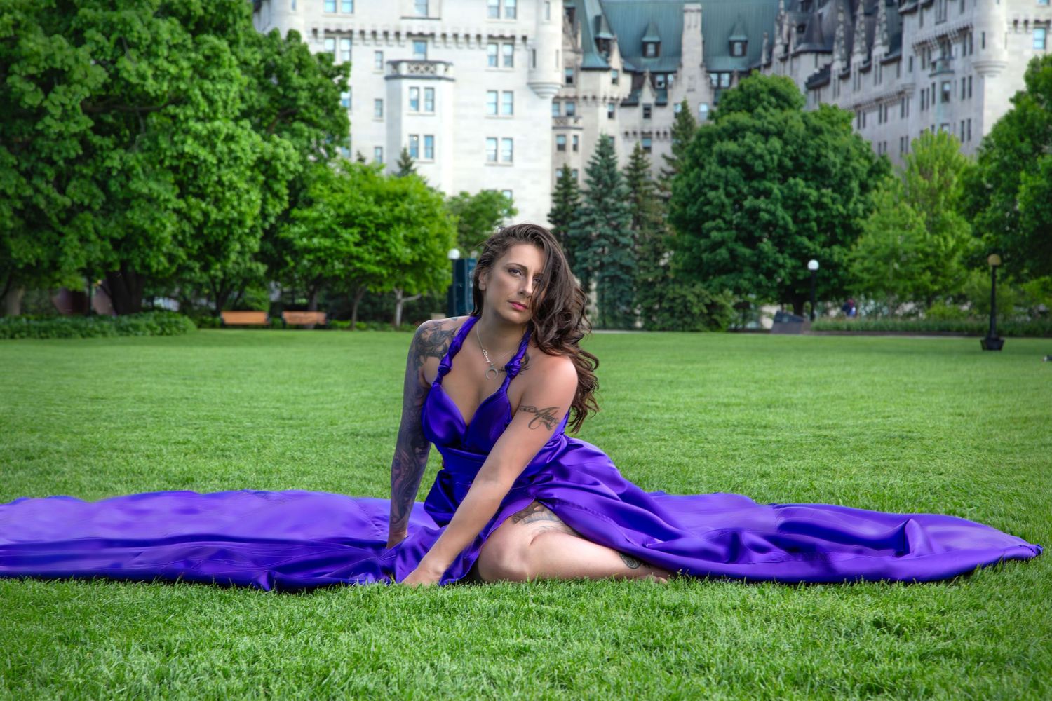 Woman in purple satin gown sits gracefully on green lawn before a grand historic building. Ottawa flying dress luxury photography. Cheateau