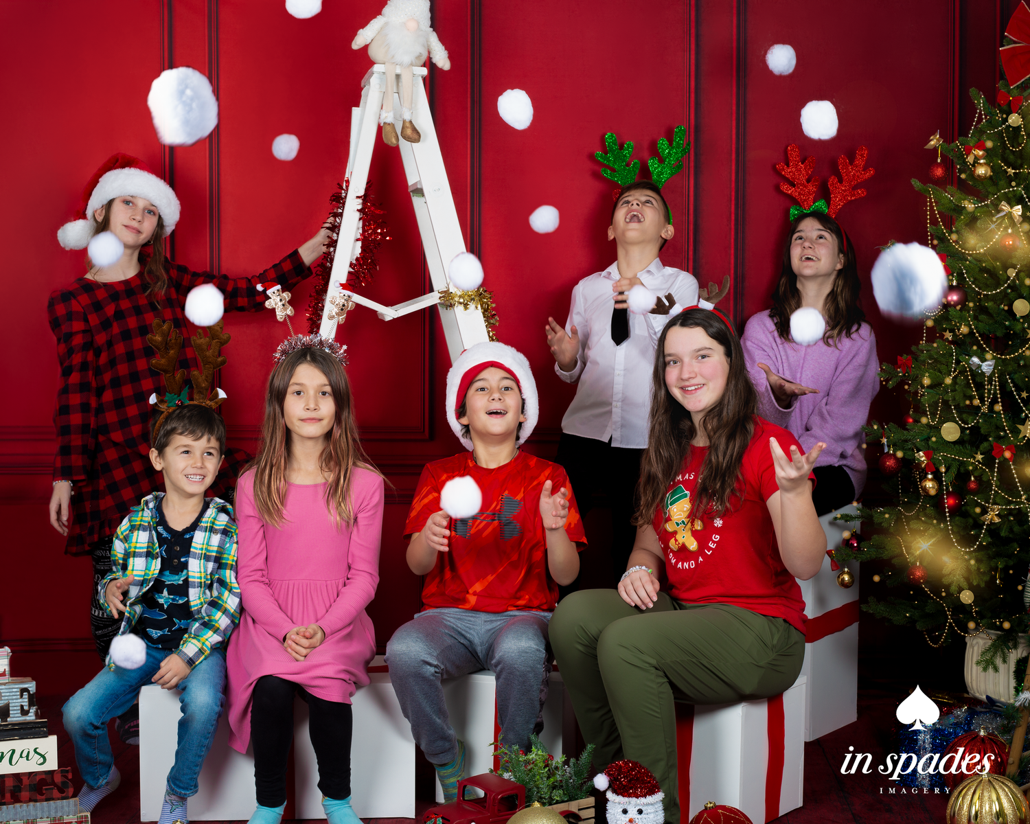 Seven children in festive Christmas attire pose playfully with snowballs in front of a red backdrop with holiday decorations.
