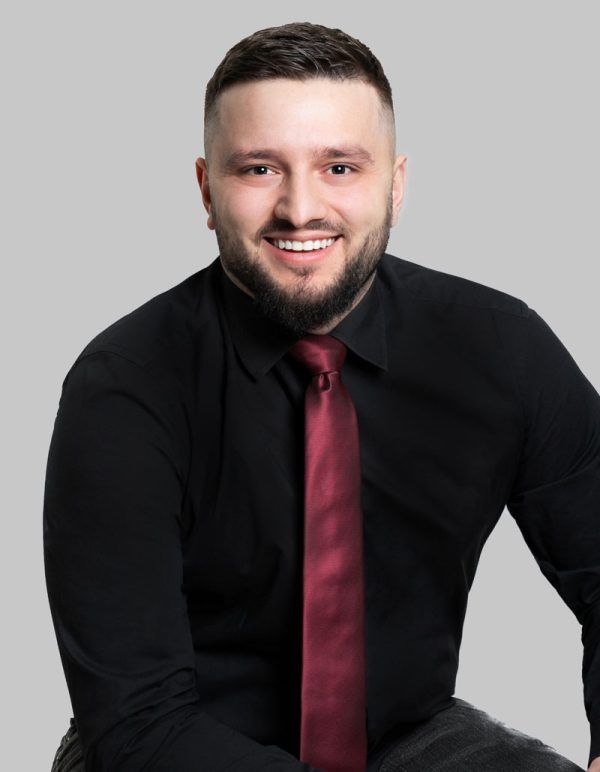 Smiling man with beard in black shirt and burgundy tie against gray background.