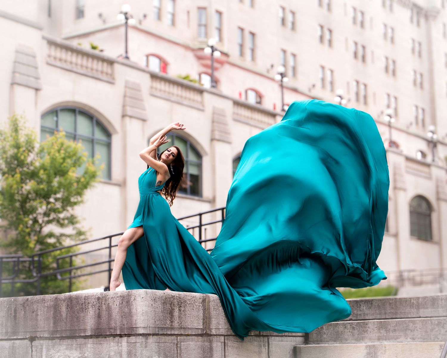 Woman in dramatic flowing teal gown poses on stone wall before urban building. Ottawa flying dress luxury photography. Château Laurier