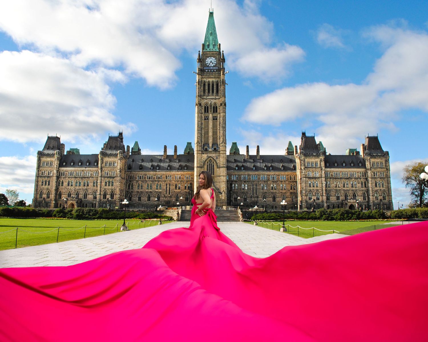 Woman in vibrant pink gown with billowing train stands before Canada's Parliament Hill building. Ottawa flying dress photography.