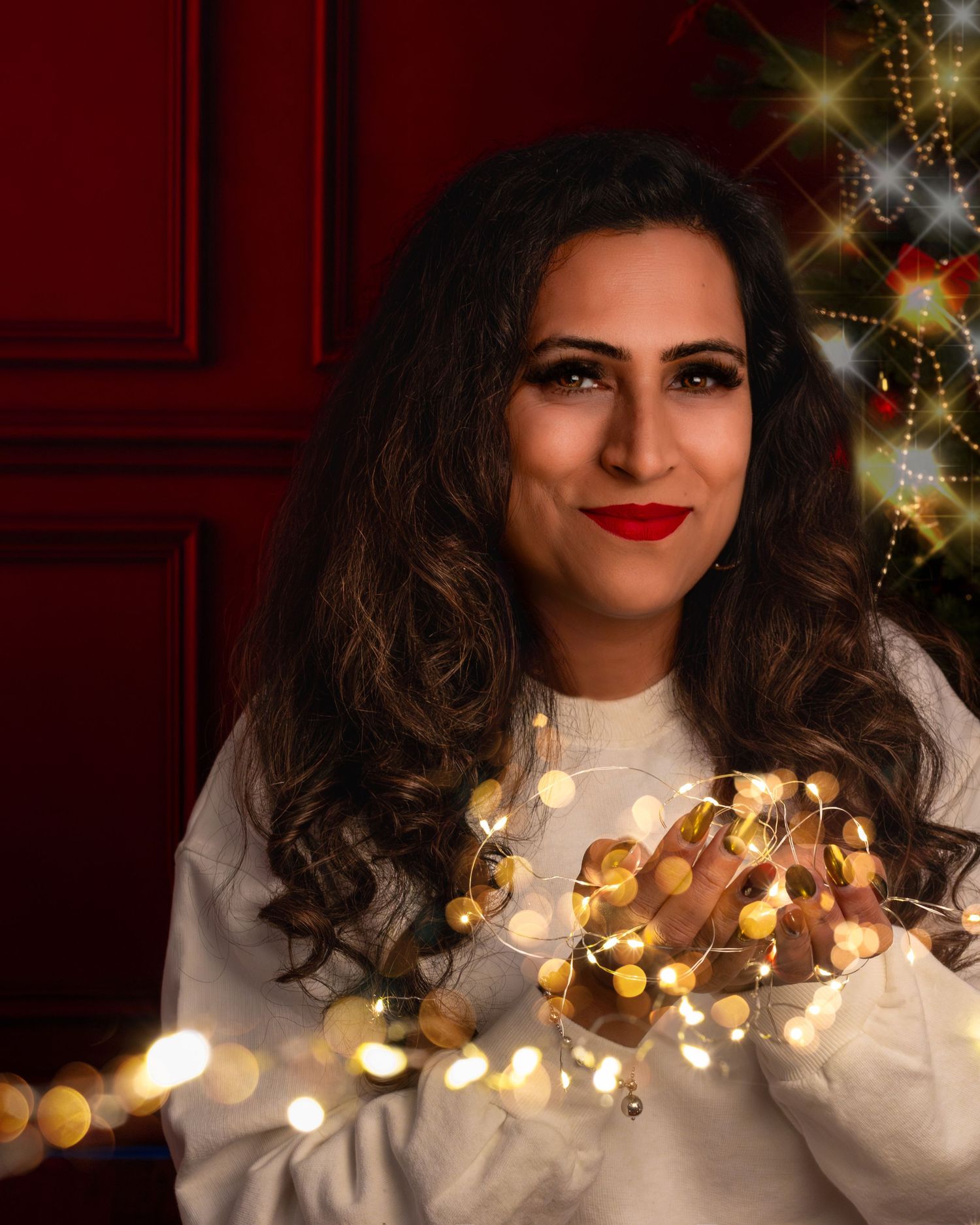 Woman with red lips holding glowing fairy lights near a decorated Christmas tree with a red door background.