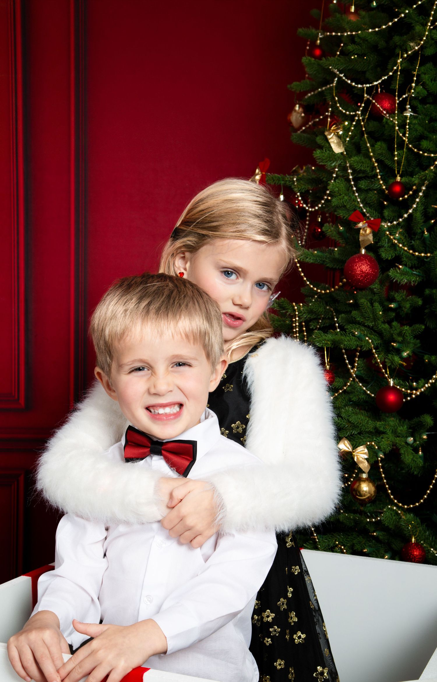 Two blonde children in festive attire pose by a decorated Christmas tree, boy in bow tie, girl in white fur wrap.
