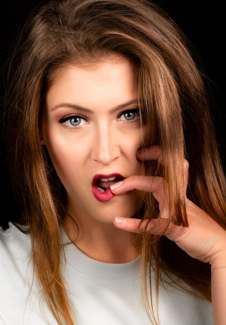 Young woman with blue eyes and red lips poses with finger near mouth on dark background.