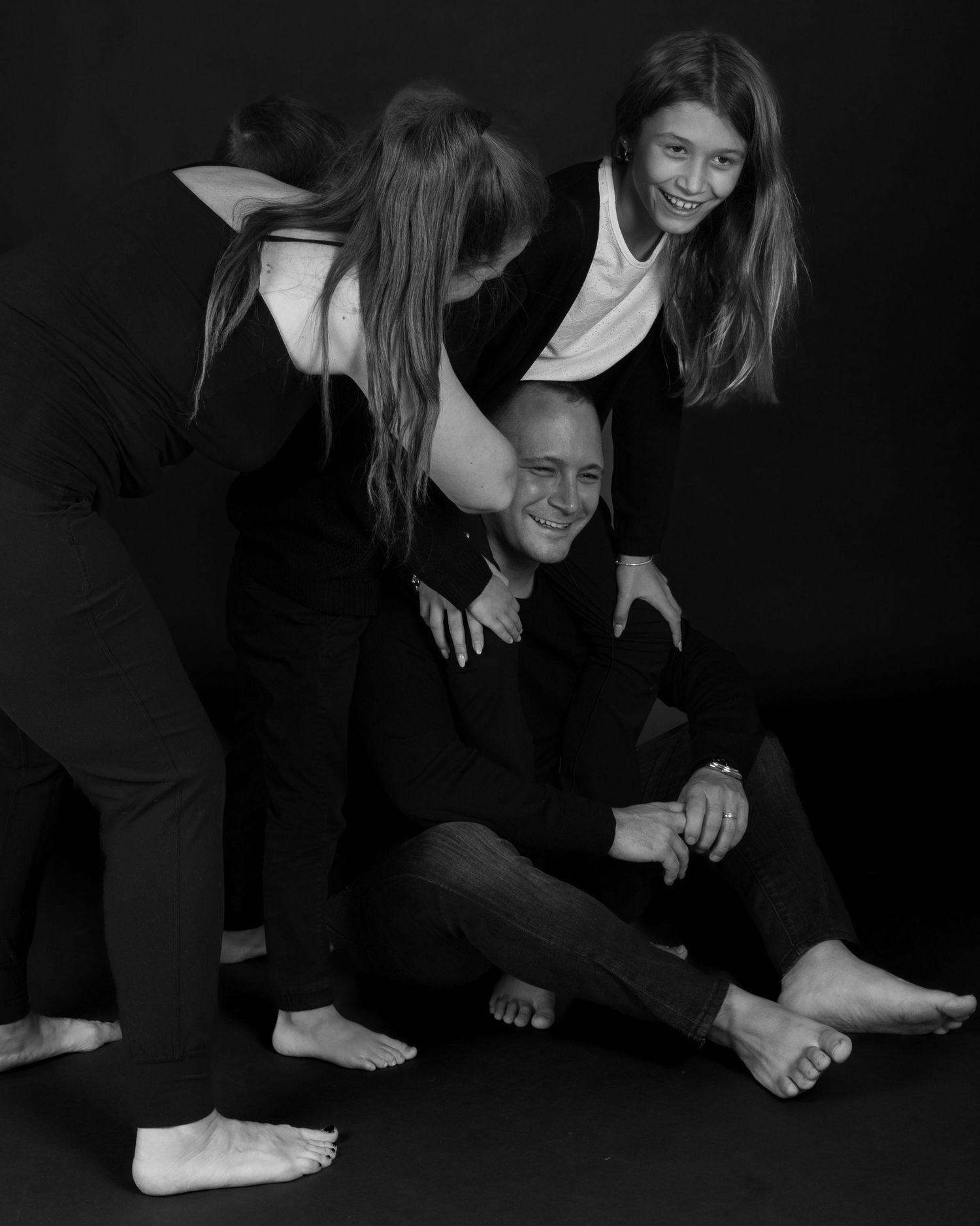Black and white family portrait with man sitting on floor while two women lean over him laughing. Behind the scenes BTS