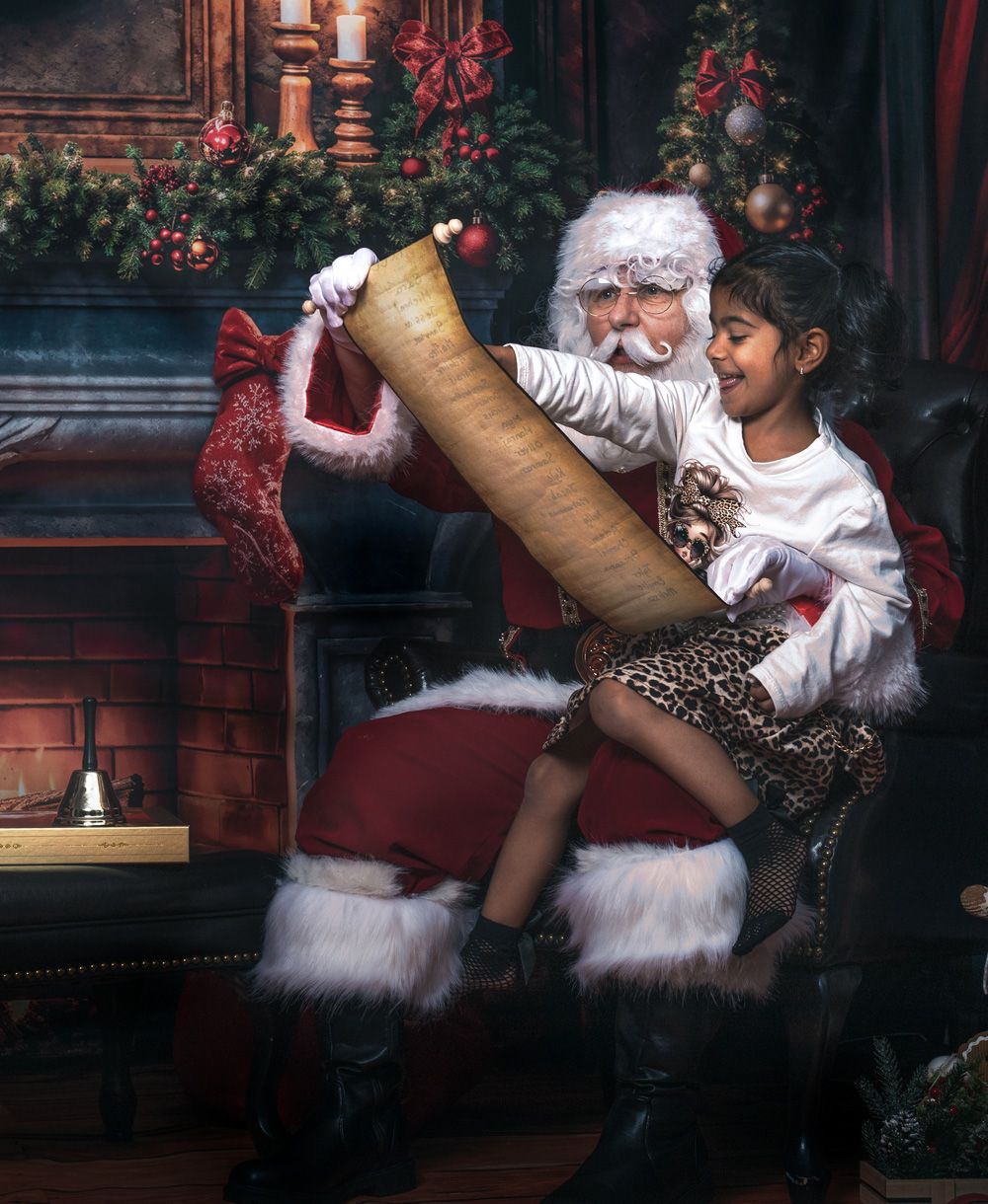A young girl sits on Santa's lap by a fireplace, holding a large scroll with Christmas decorations in the background.