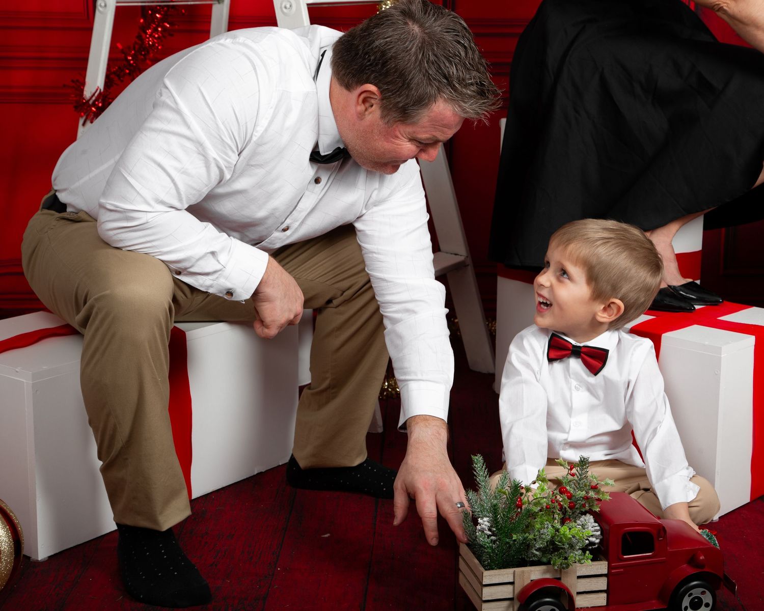 A father in a white shirt and bow tie smiles at his young son during a festive Christmas photo session with gifts.