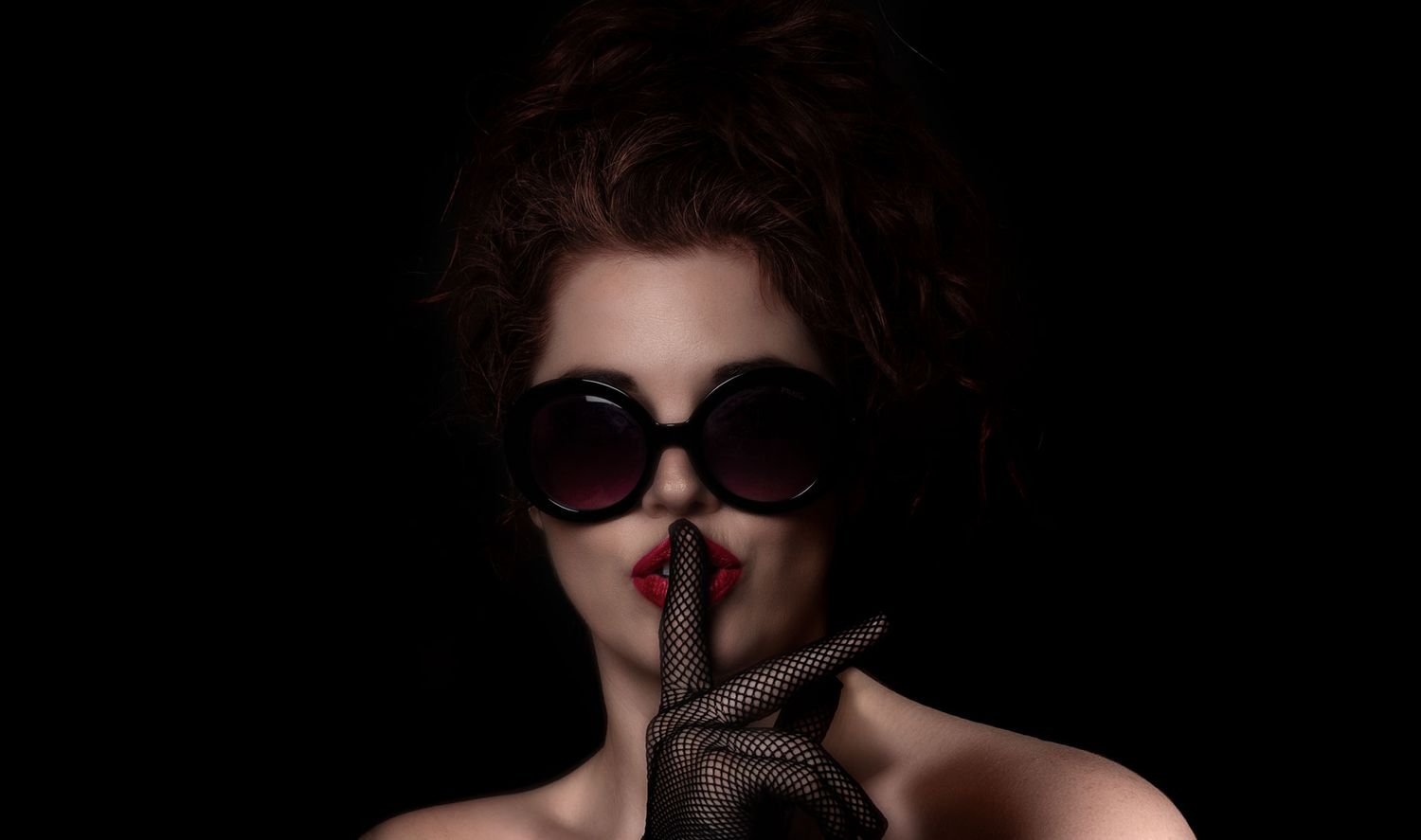 Dramatic dark portrait of a mysterious woman in sunglasses making a silence gesture with gloved finger.