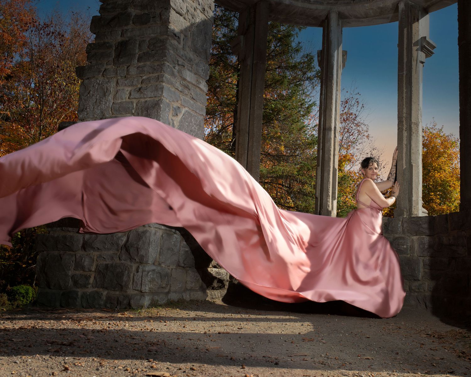 Woman in pink billowing gown stands in stone rotunda during autumn.  Ottawa flying dress photography. Mackenzie King estate