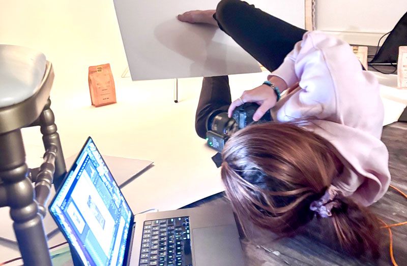 Photographer leaning over camera equipment with laptop and orange phone visible on desk during product photo shoot.