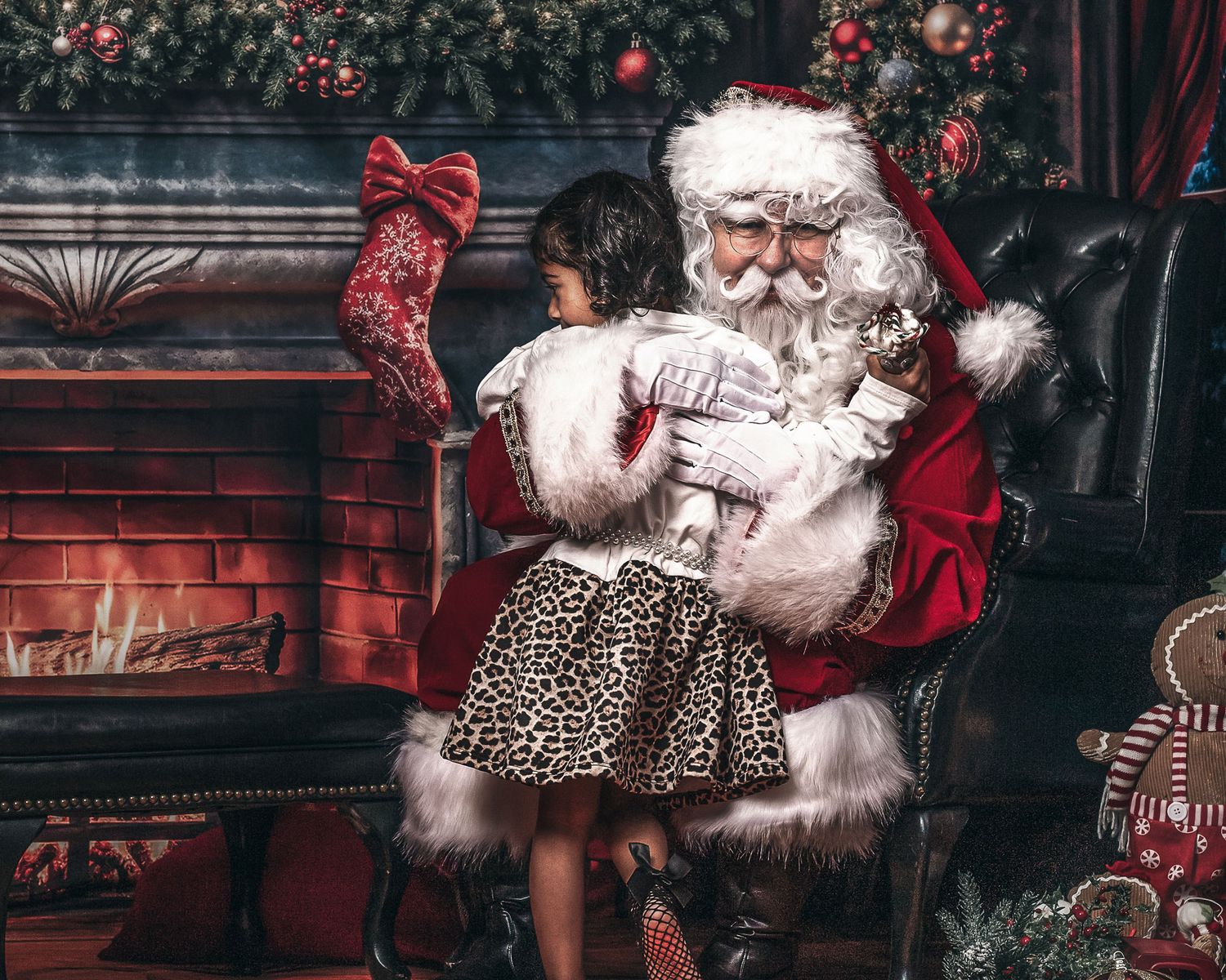 Santa Claus sits in a chair holding a young girl near a decorated fireplace with Christmas stockings and ornaments.