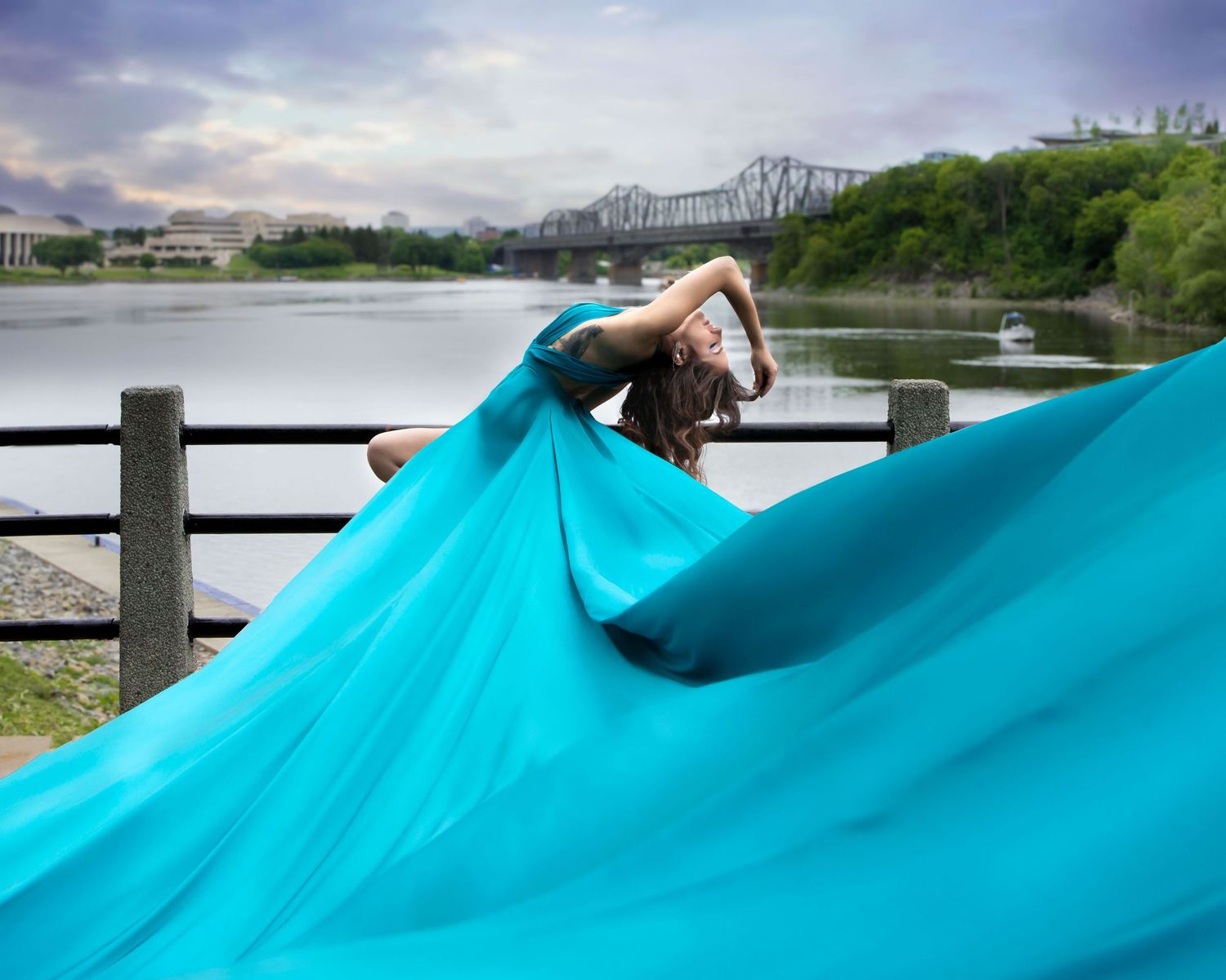 Woman in flowing teal gown poses gracefully by riverside with bridge in background. Ottawa flying dress luxury photography