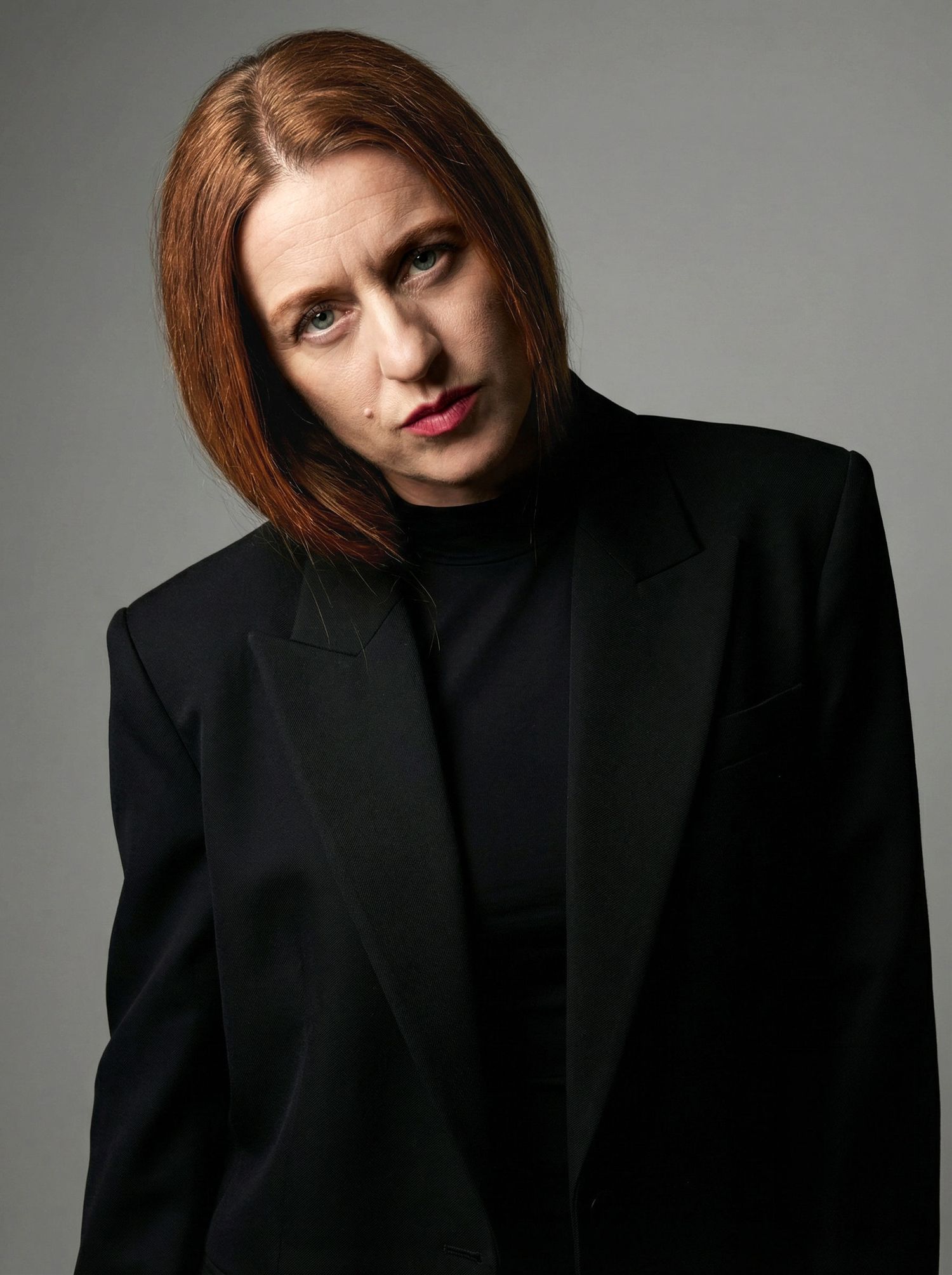 A woman with red bob hair wearing an oversized black blazer poses with a serious expression against a gray background.