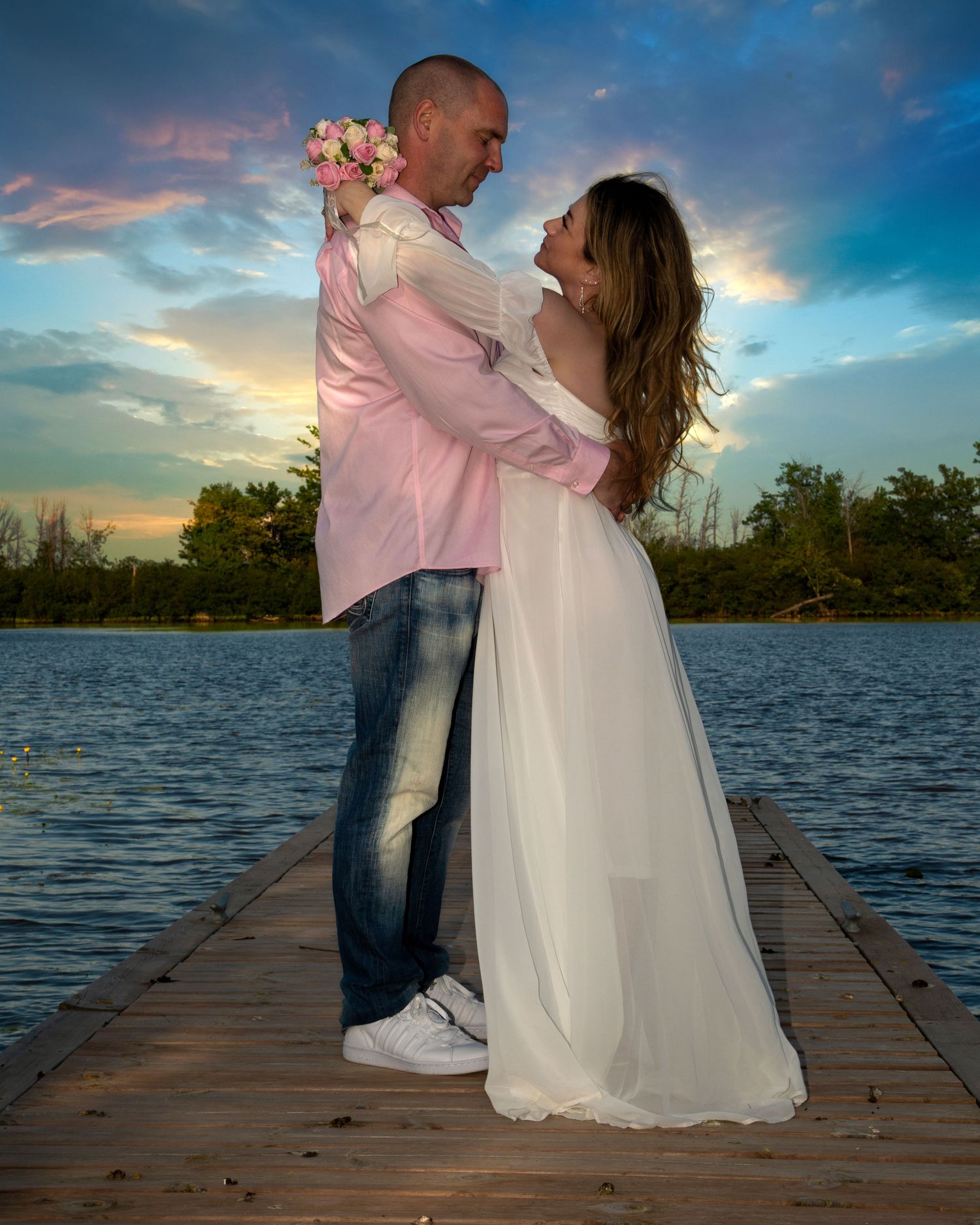 A couple in wedding attire embraces romantically on a dock at sunset with colorful sky behind them. Ottawa wedding and events