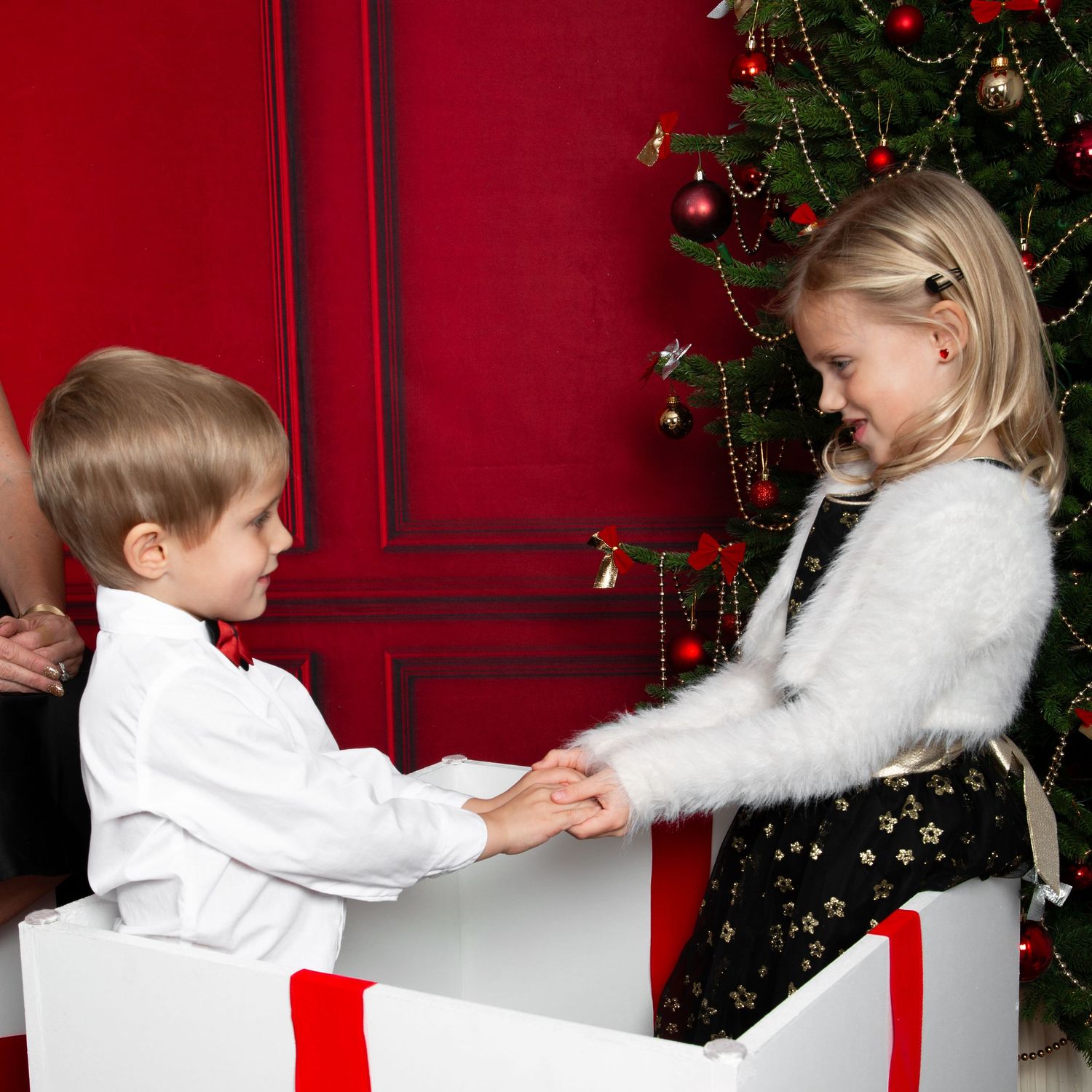 Two young children exchanging gifts near a decorated Christmas tree with red ornaments and wrapped presents.