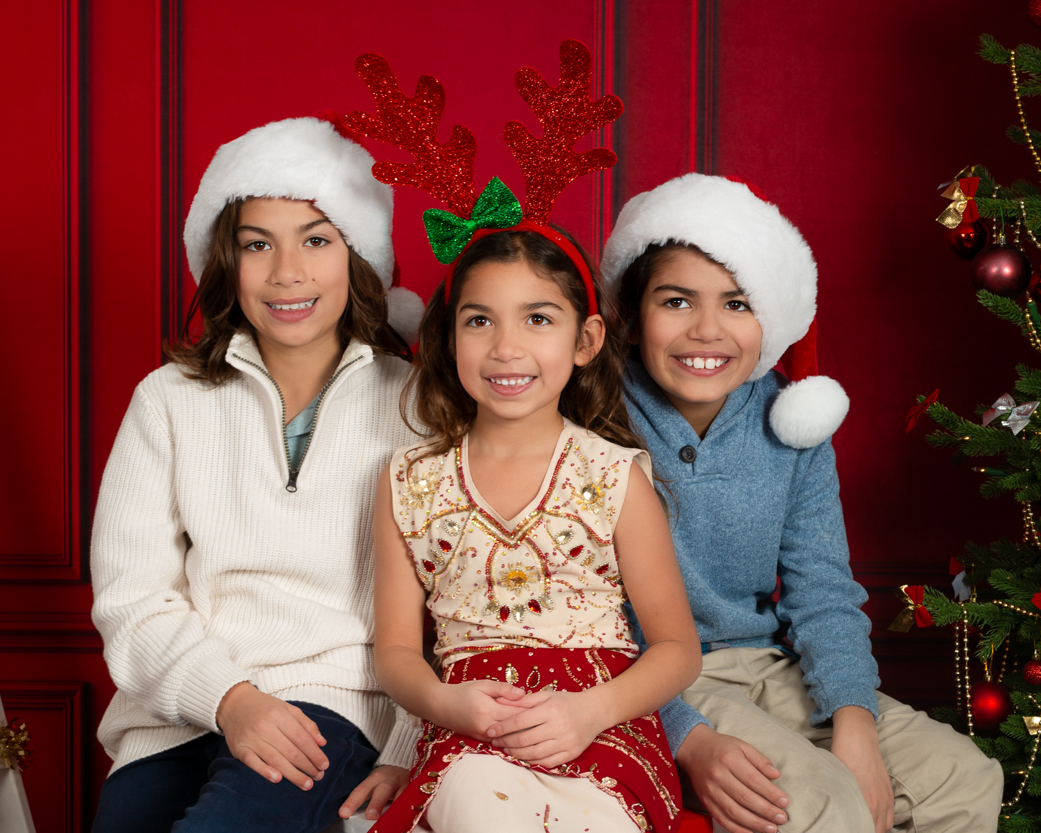 Three children in Santa hats pose together for a festive Christmas photo in front of a red background.