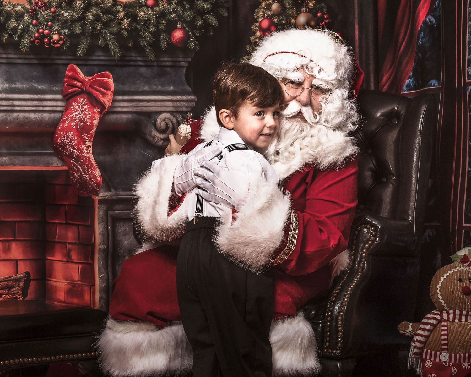 A young boy sits on Santa Claus's lap near a decorated fireplace with stockings during a Christmas photo session.