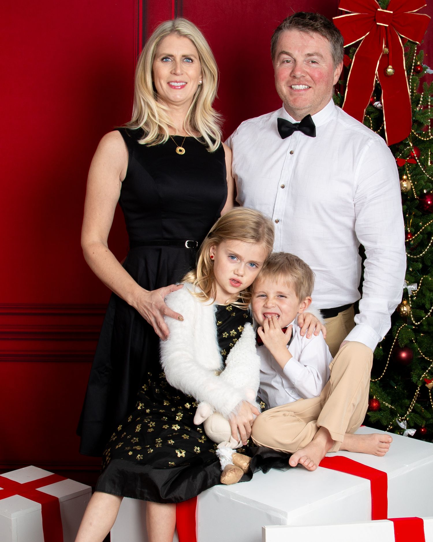 Family of four posing for a festive Christmas portrait in front of a decorated tree with wrapped gifts.